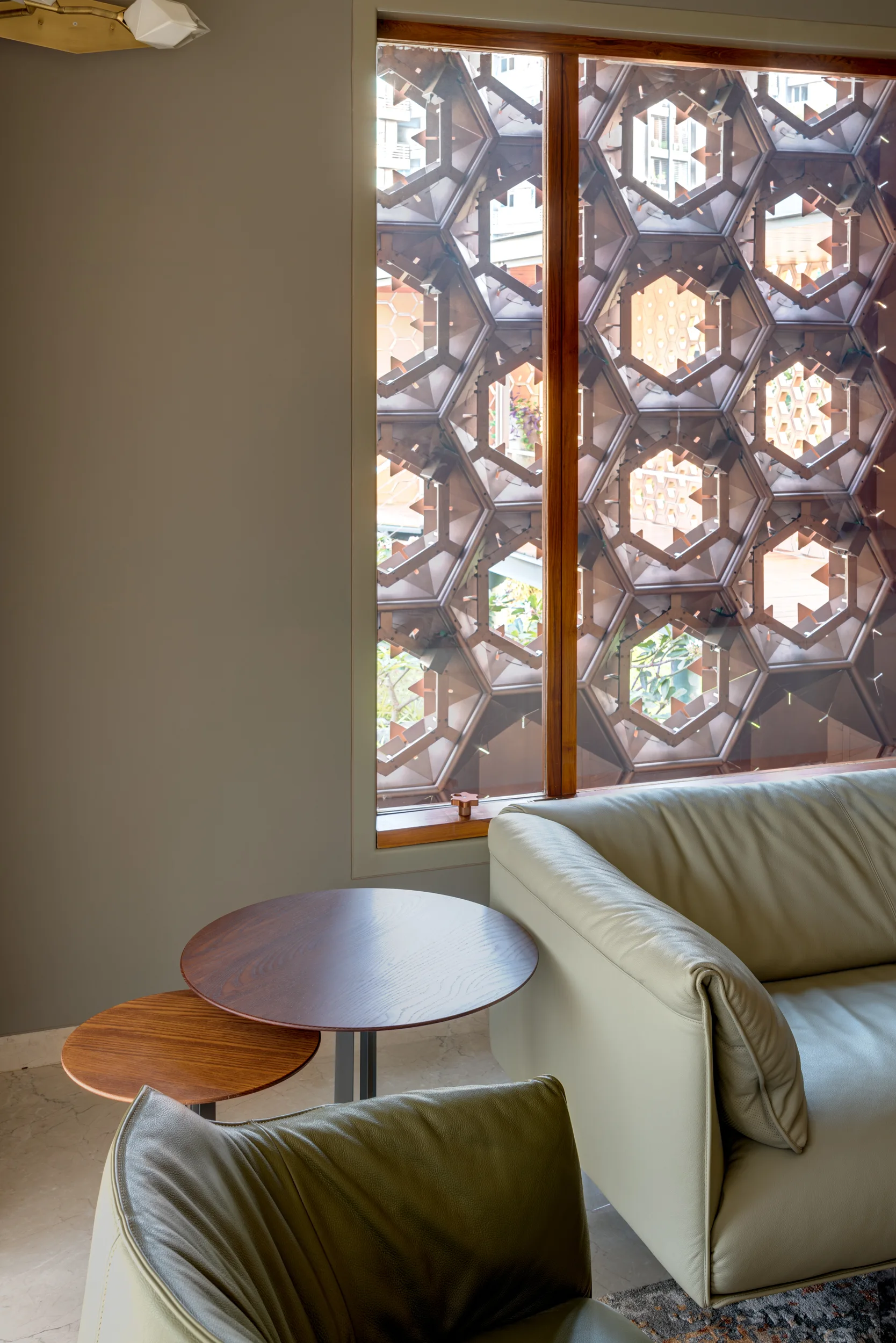 A modern grey living room features a leather sofa next to tiered wooden side tables and a window with a bronze geometric pattern screen.
