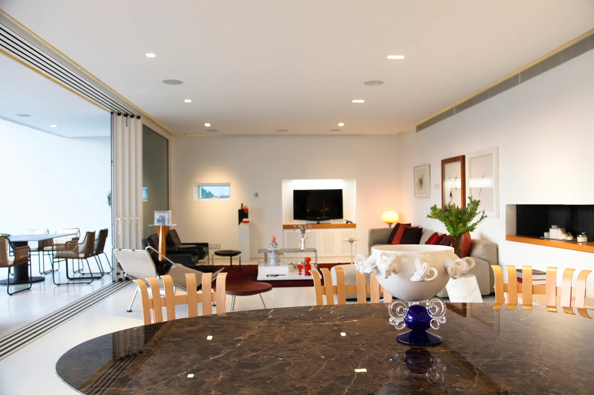 A polished brown marble dining table with a decorative bowl sits in the foreground of a bright open-plan living room with white walls and folding glass doors.