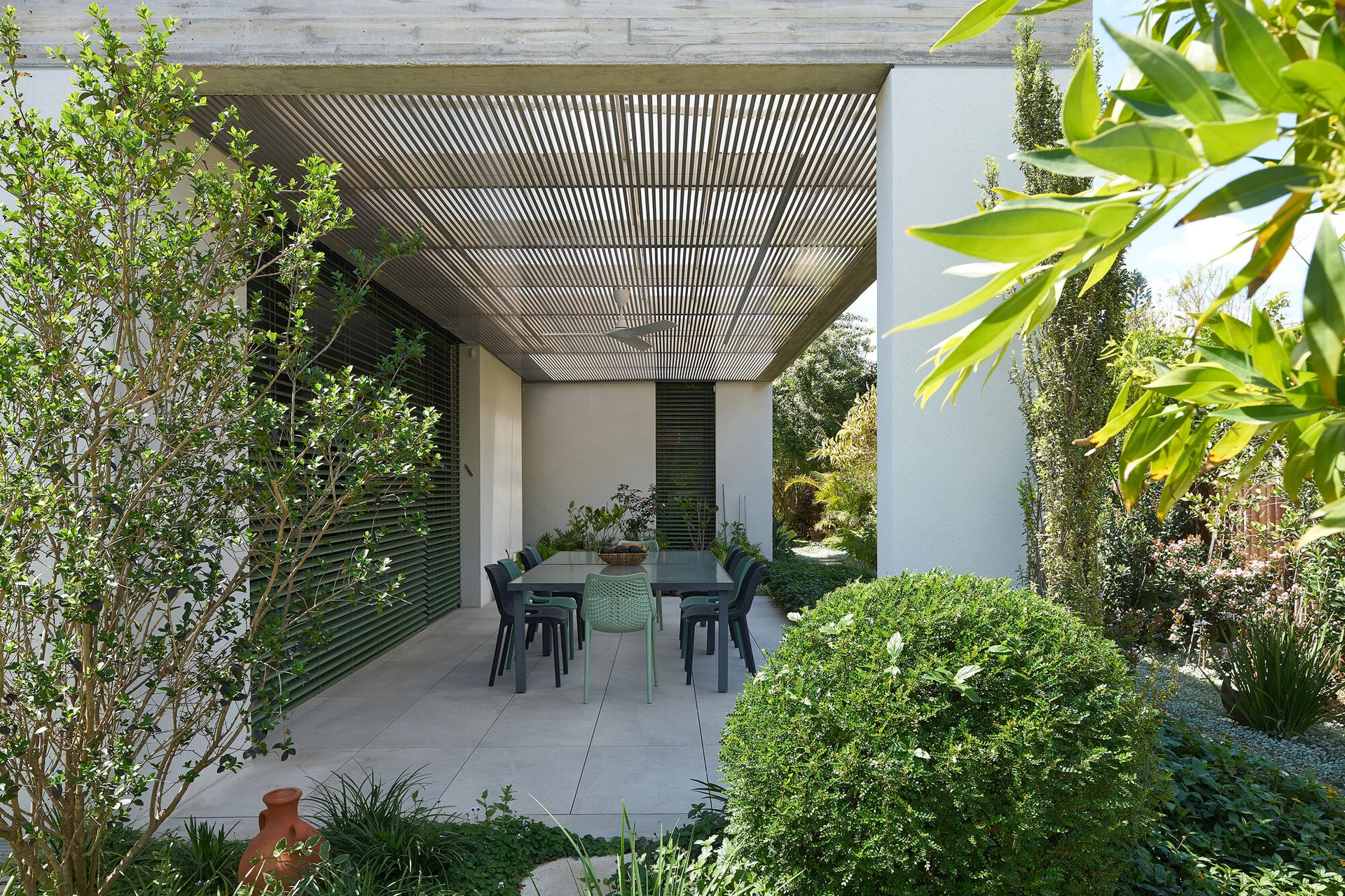 A covered outdoor patio featuring an outdoor dining table with green and dark chairs beneath a slatted ceiling surrounded by a lush green garden.
