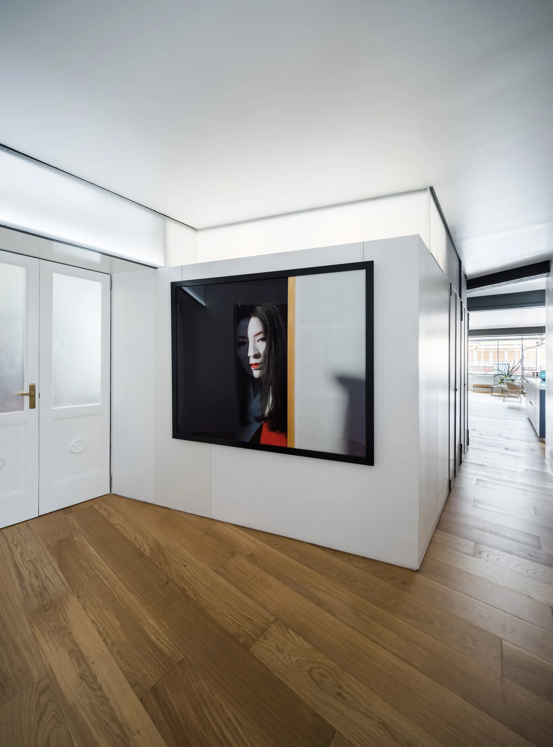 A white minimalist hallway features a central partition wall with a large framed portrait photograph over warm hardwood flooring.