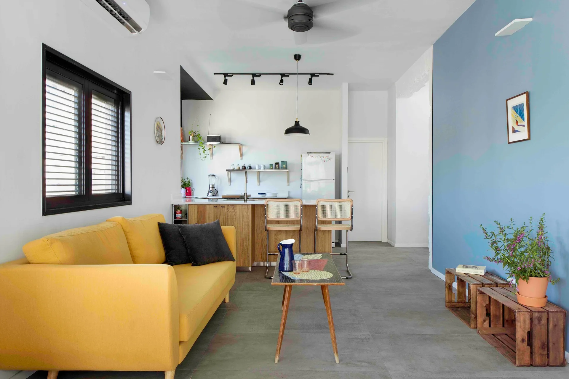A modern open-plan living area featuring a yellow sofa, a blue accent wall, and a wood-toned kitchen with cane bar stools.
