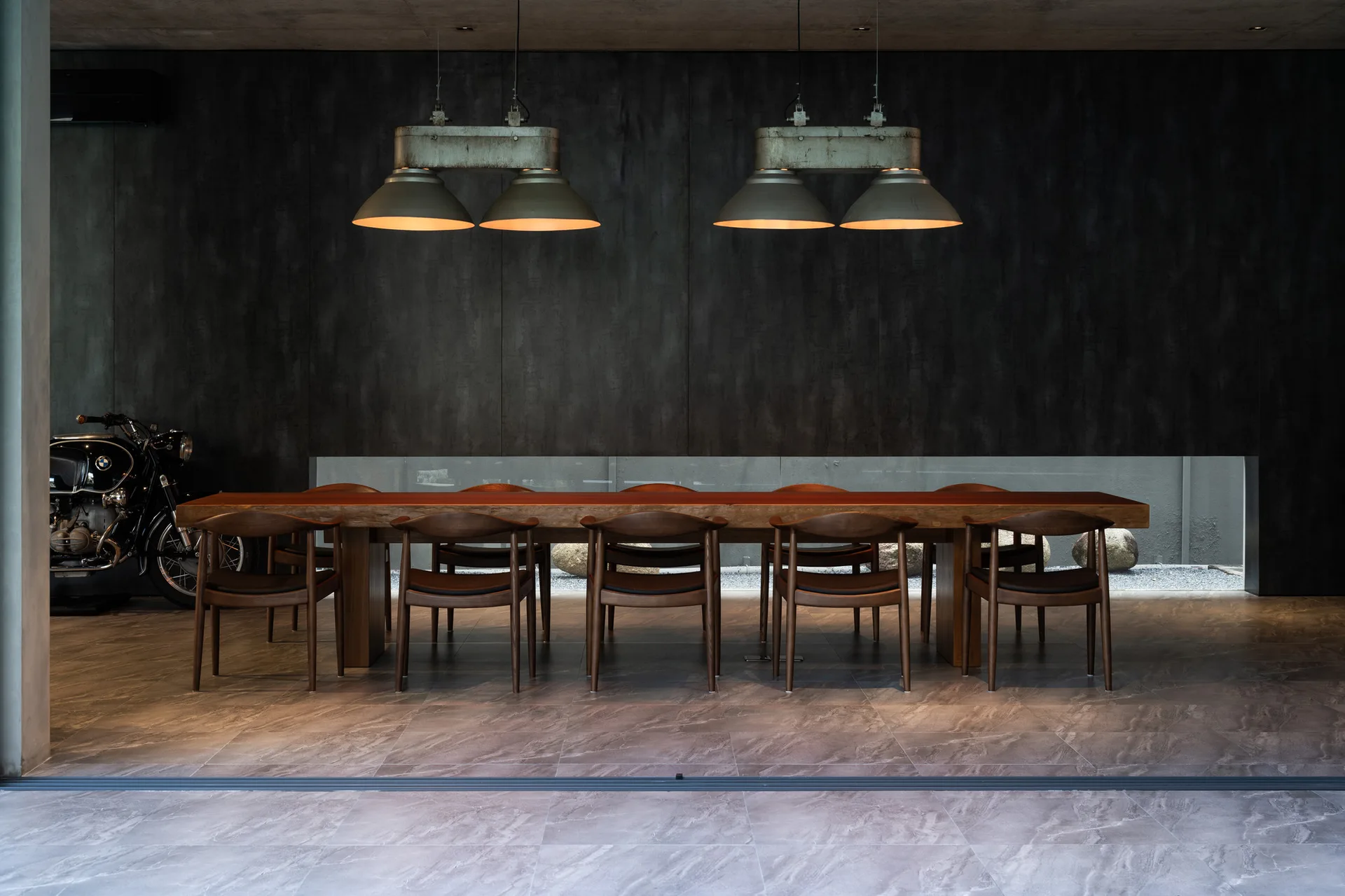 A long wooden dining table and chairs in a moody industrial dining room with a dark wall, pendant lights, and a low horizontal window.