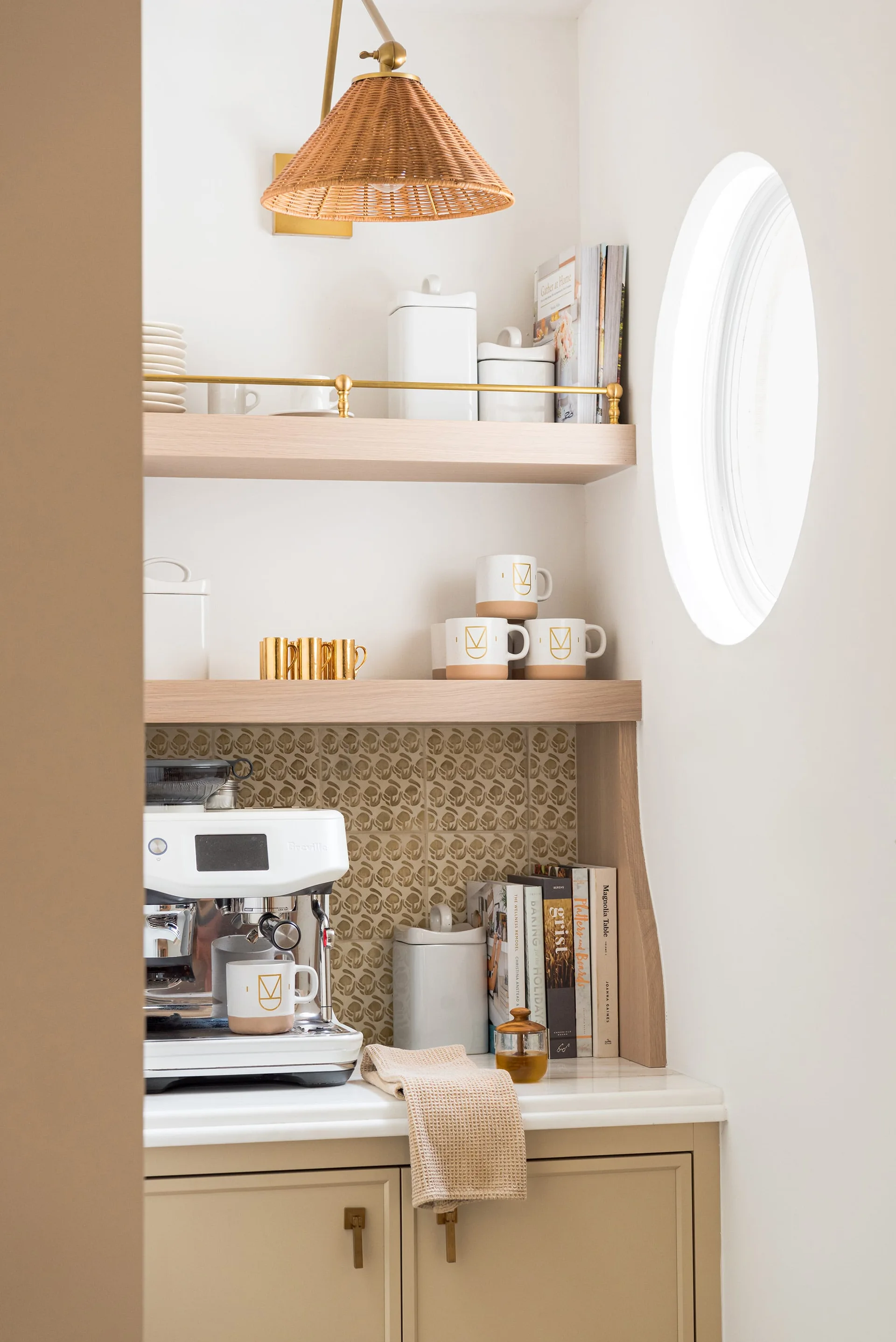 A kitchen coffee nook featuring a white espresso machine on a marble counter, wooden shelves, a patterned backsplash, and a round wall window.