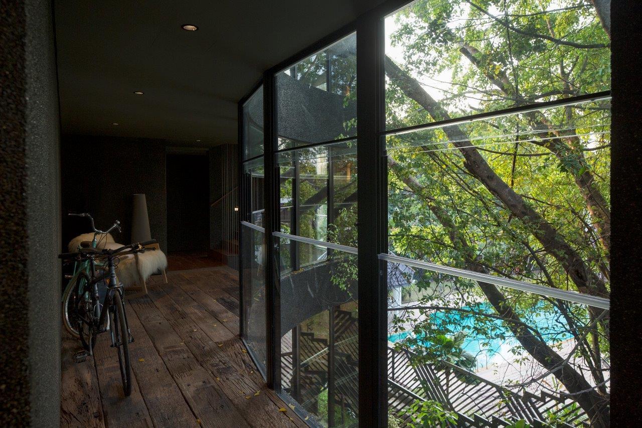 A dark hallway features rustic wooden plank flooring, a floor-to-ceiling glass window wall overlooking green trees, a bicycle, and a fur-covered bench.