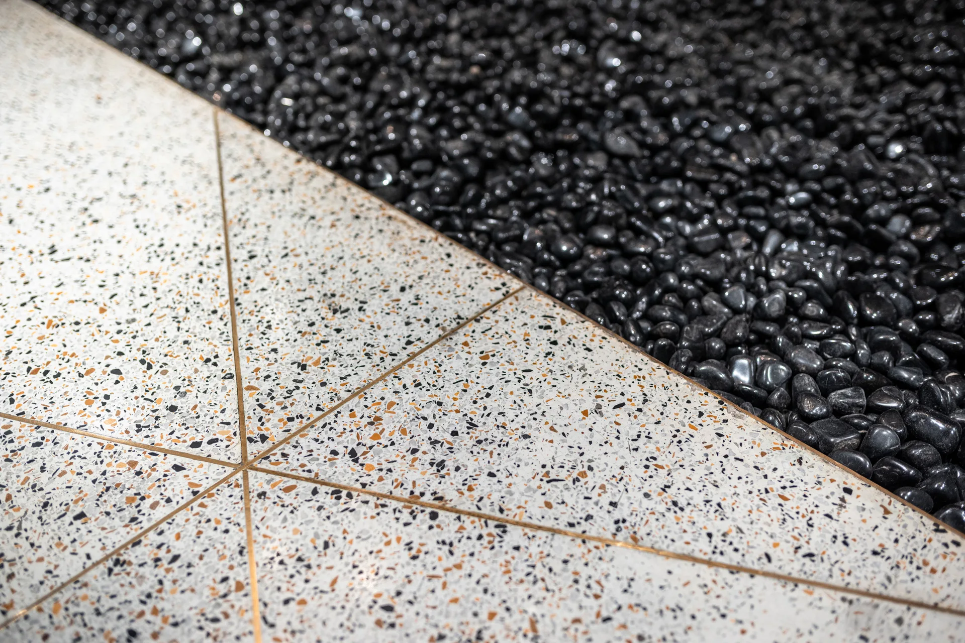 A close-up view of polished white terrazzo floor tiles with geometric brass inlays meeting a border of glossy black pebbles.