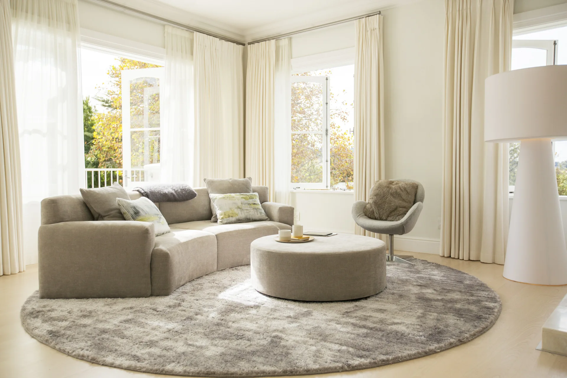 A curved beige sofa and a round ottoman sit on a circular grey rug in a bright room with floor-to-ceiling curtains.