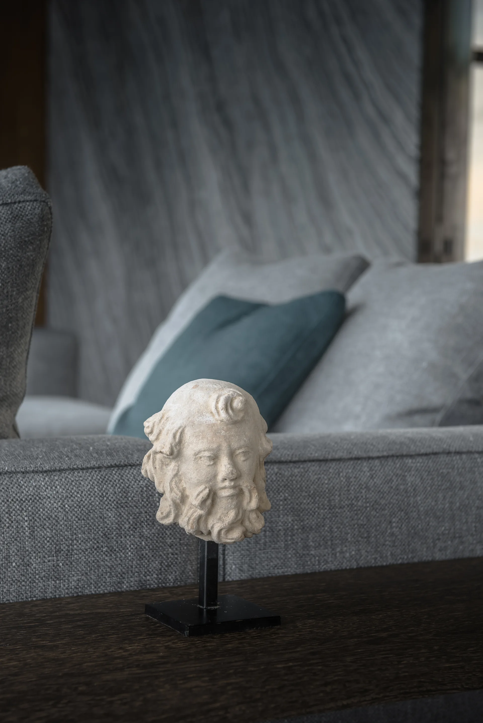 A stone-like sculpted bust on a black stand sits on a dark wood console table with a grey sofa in the background.