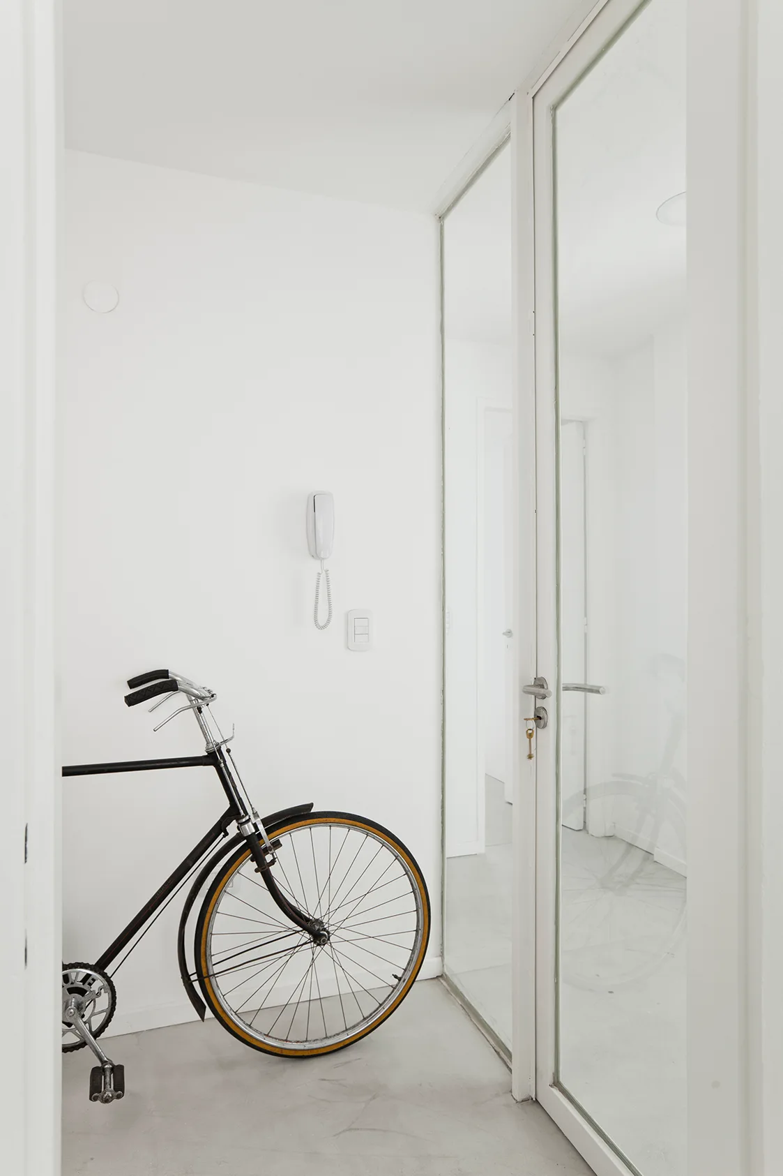 A black bicycle stands parked on a light gray floor in a minimalist white entryway next to a tall glass-paned door.