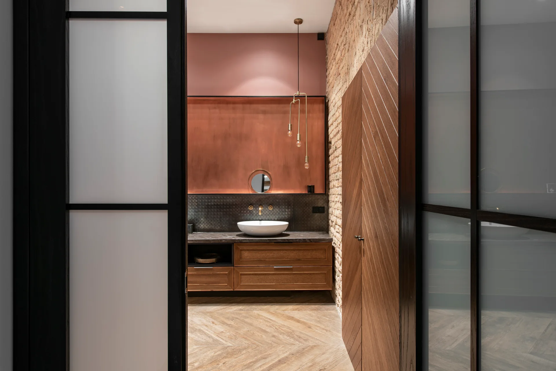 A modern bathroom interior featuring a wooden vanity, copper backsplash, white vessel sink, and an exposed brick wall seen through a glass door.