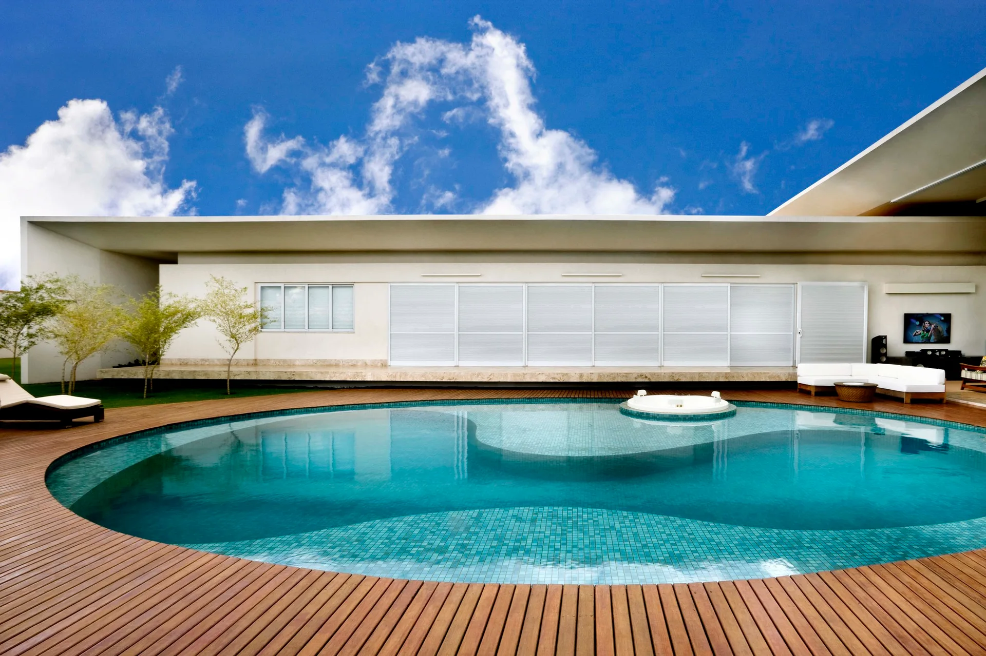 A curved blue swimming pool is surrounded by a natural wood deck next to a minimalist white house with sliding window panels.