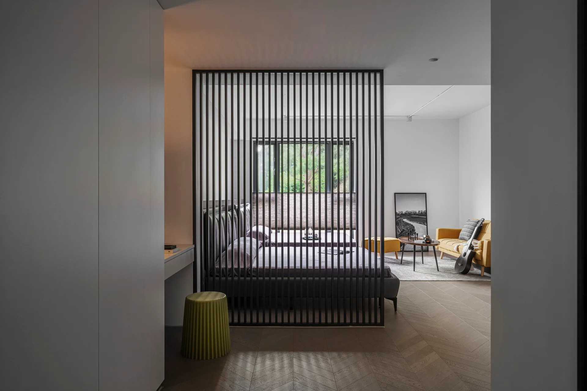A bedroom with an upholstered bed is partitioned from a living area with a yellow sofa by a tall black slatted metal screen.