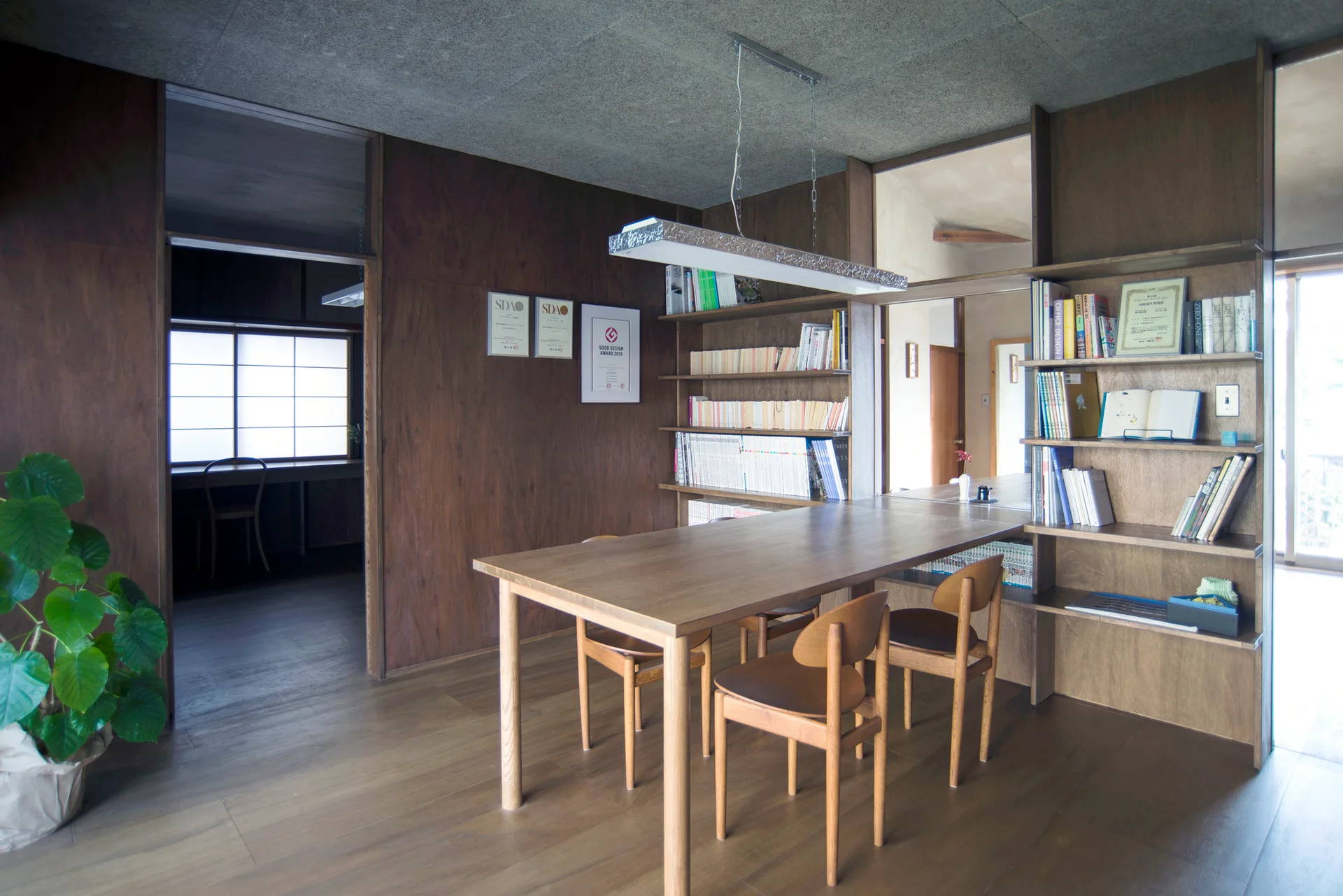 A wooden dining table and chairs sit in a room with dark wood paneled walls, built-in bookshelves, and a textured grey ceiling.