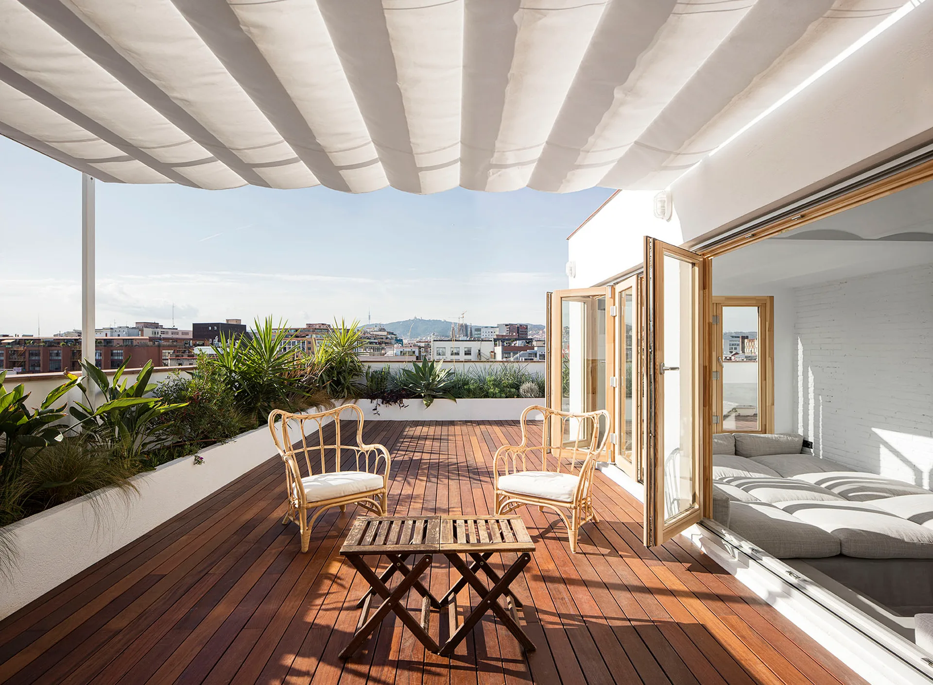 A rooftop terrace features dark wood decking, rattan lounge chairs under a white fabric canopy, and open folding glass doors leading indoors.