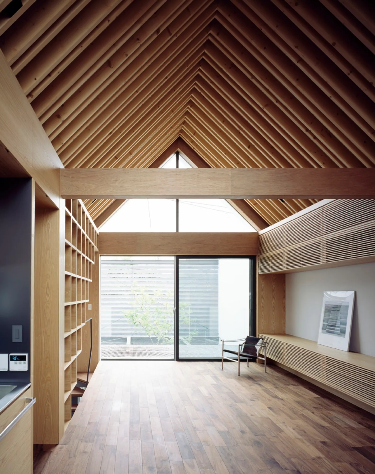 A minimalist living space features a high vaulted wood-slatted ceiling, custom timber shelving, and a sliding glass door.
