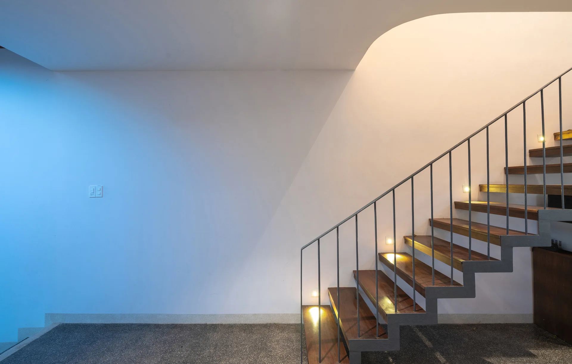 A minimalist staircase features floating wood treads, a slender metal railing, and small square built-in wall lights leading up to a curved ceiling.