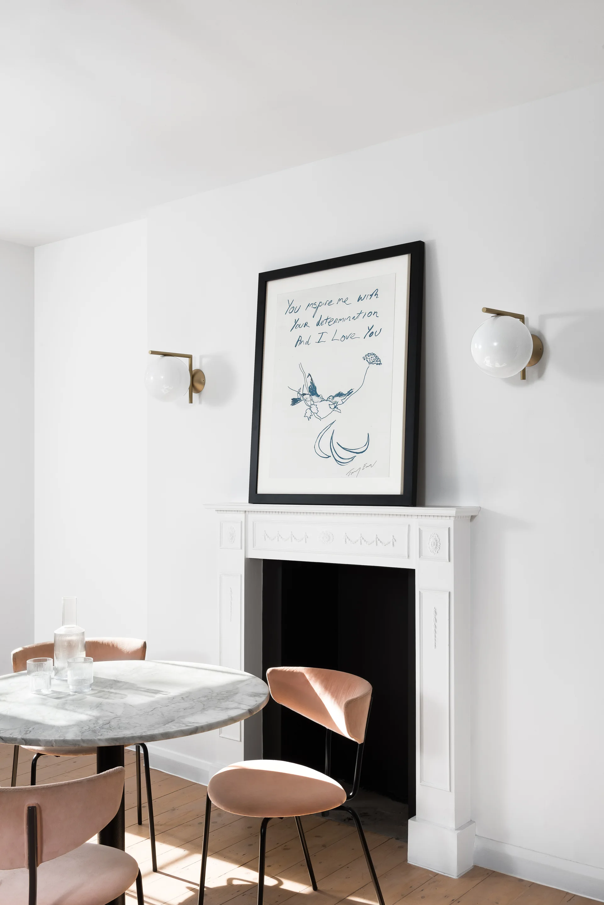 A round marble dining table with pink chairs sits next to a white fireplace topped with a framed artwork and brass globe sconces.