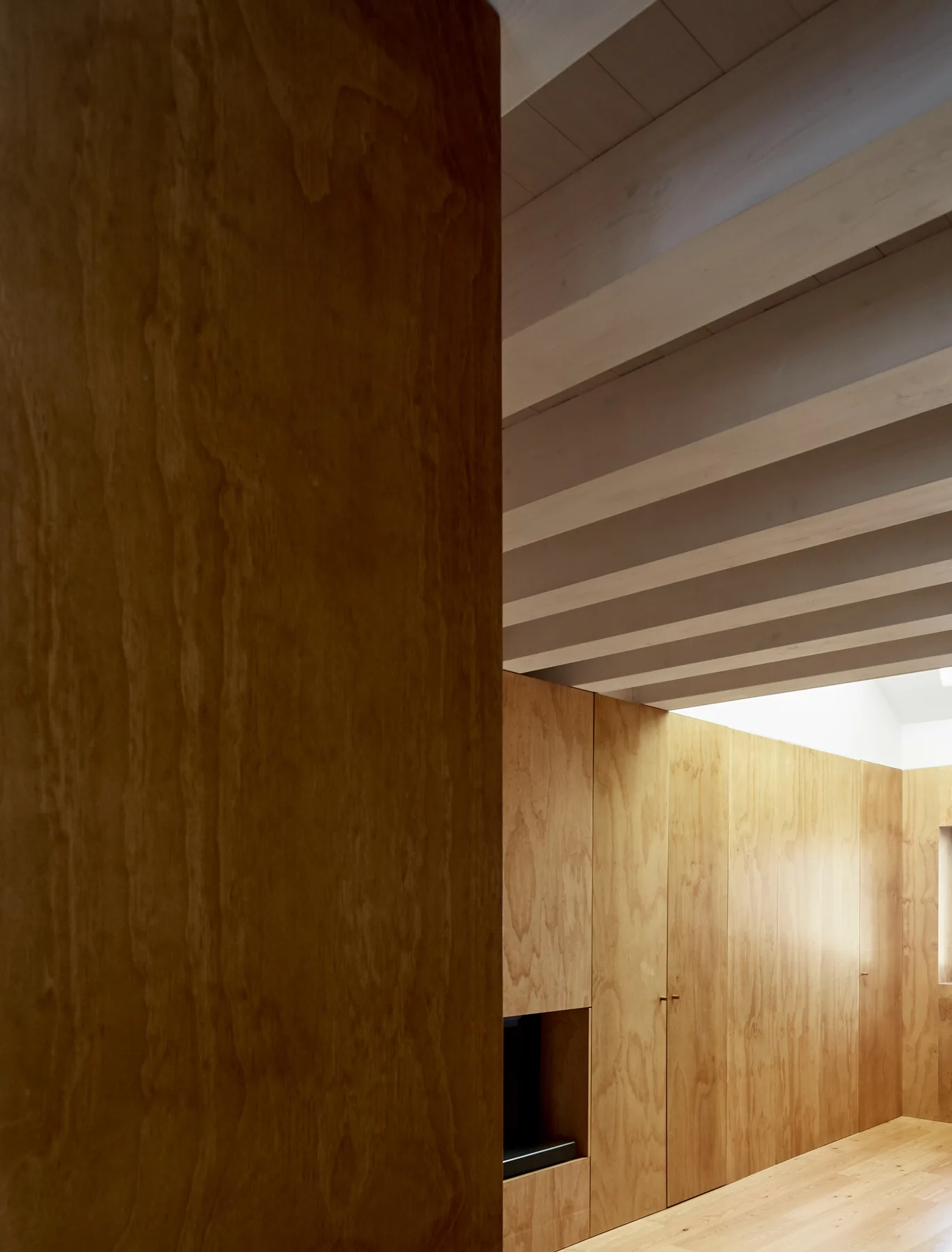 An interior space featuring warm plywood wall paneling, built-in cabinets, and exposed pale wood ceiling beams above a matching floor.