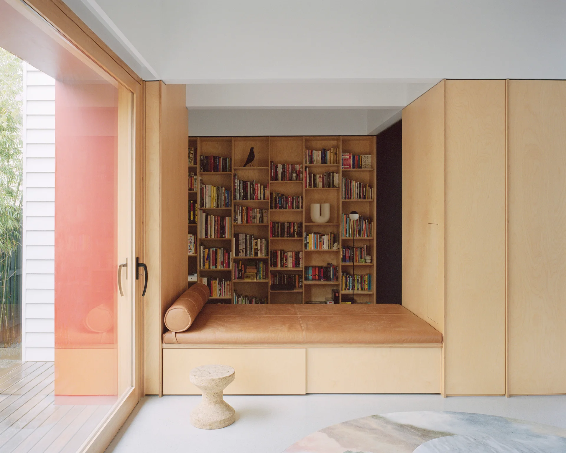 A light wood built-in shelving unit with a tan leather cushion daybed next to a sliding glass door in a minimalist room.