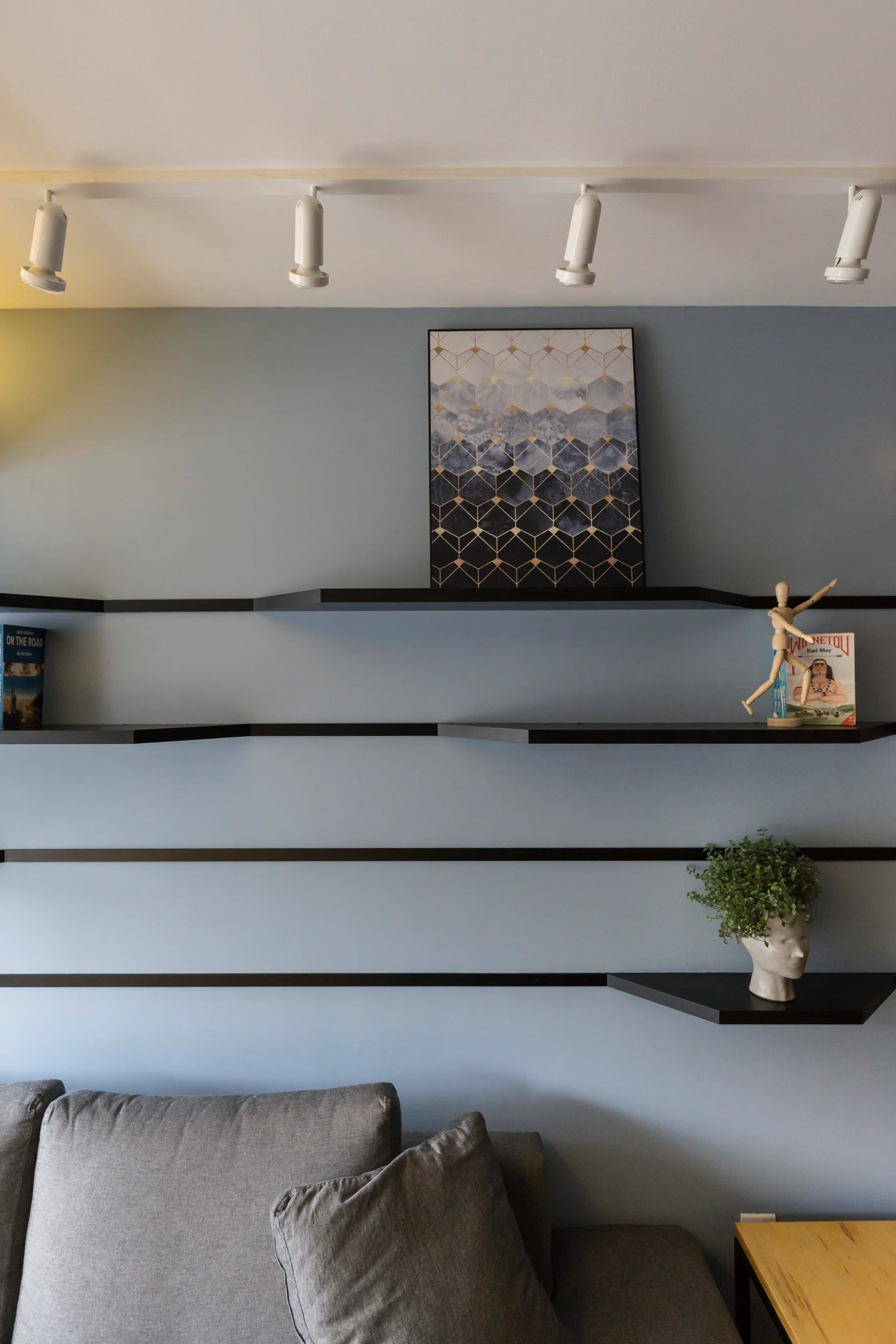 A gray fabric sofa sits below minimalist black floating shelves mounted on a dusty blue wall, styled with a geometric art piece and small decor.