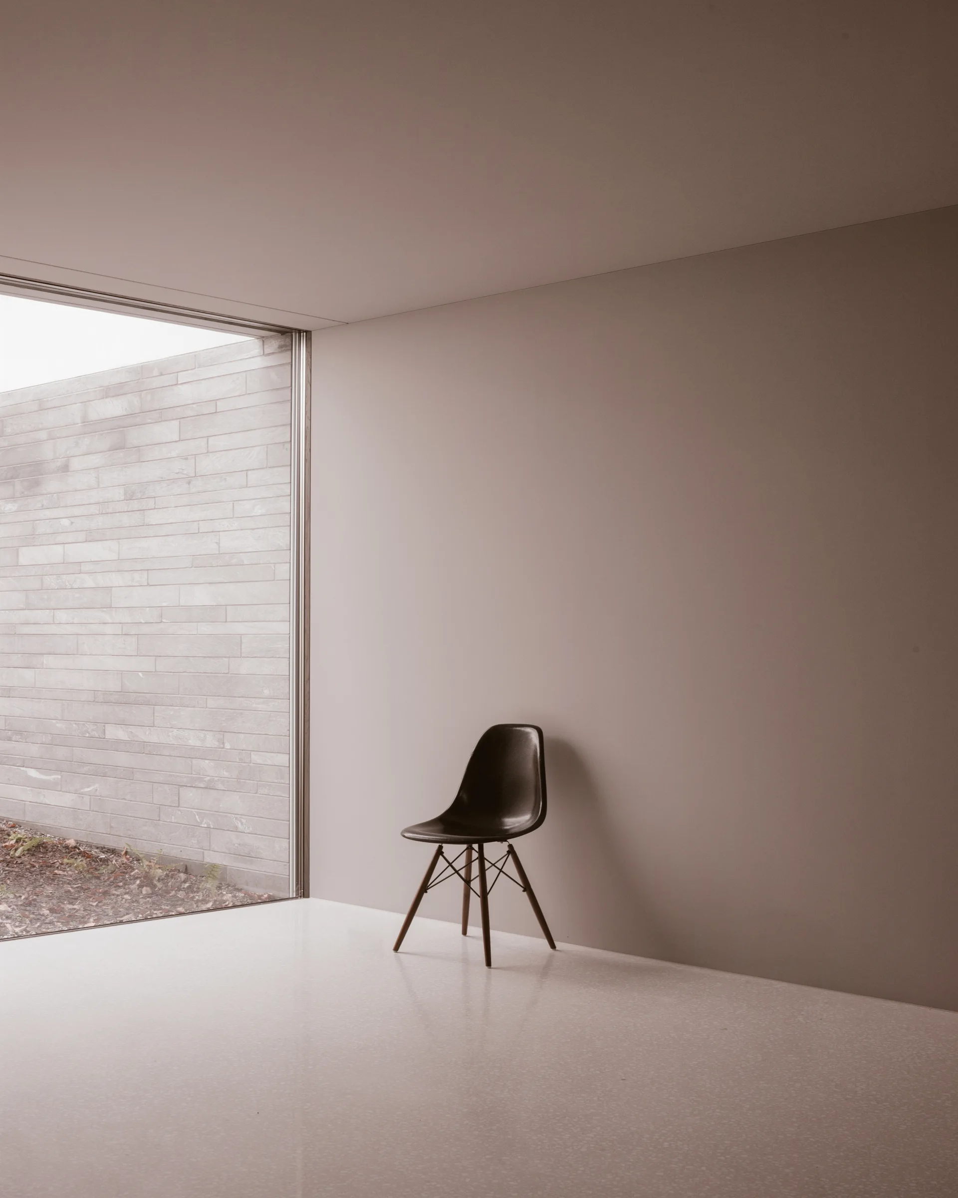 A dark molded chair stands in a minimalist room with warm beige walls next to a large floor-to-ceiling window looking out at a stone wall.