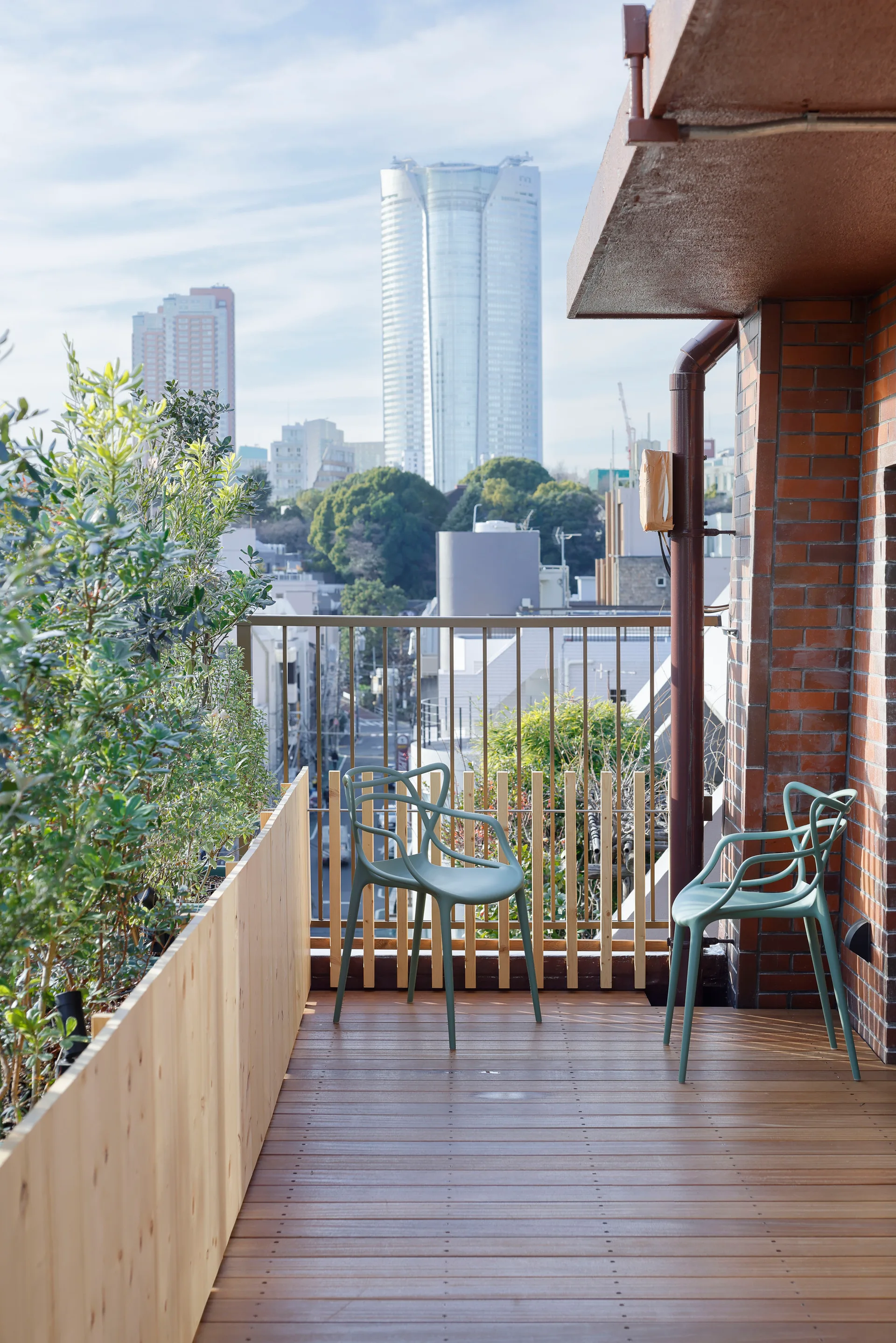 Two sage green plastic chairs sit on a wood-decked balcony next to a wooden planter box and a red brick wall with city views.
