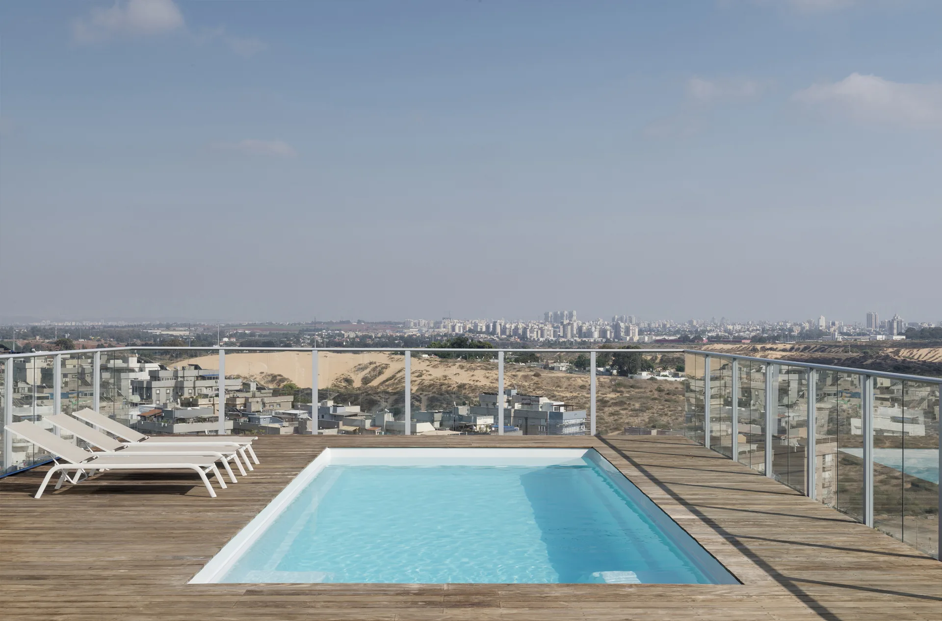 A rectangular swimming pool is built into a weathered wooden rooftop deck equipped with three white lounge chairs and a glass safety railing.