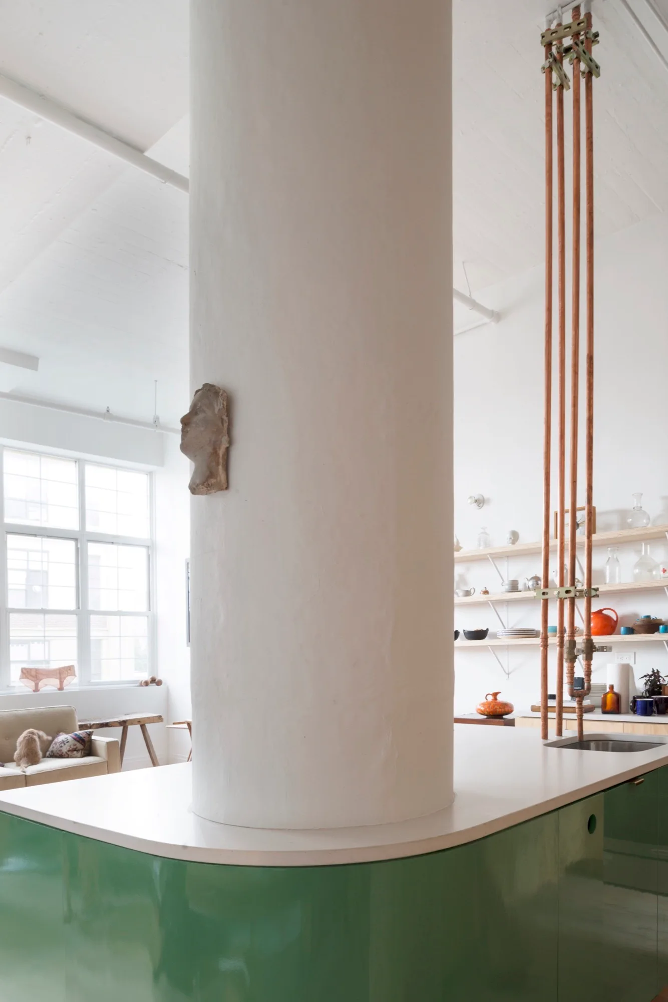 A bright industrial loft kitchen featuring a glossy green island integrated around a large white column and exposed vertical copper pipes.