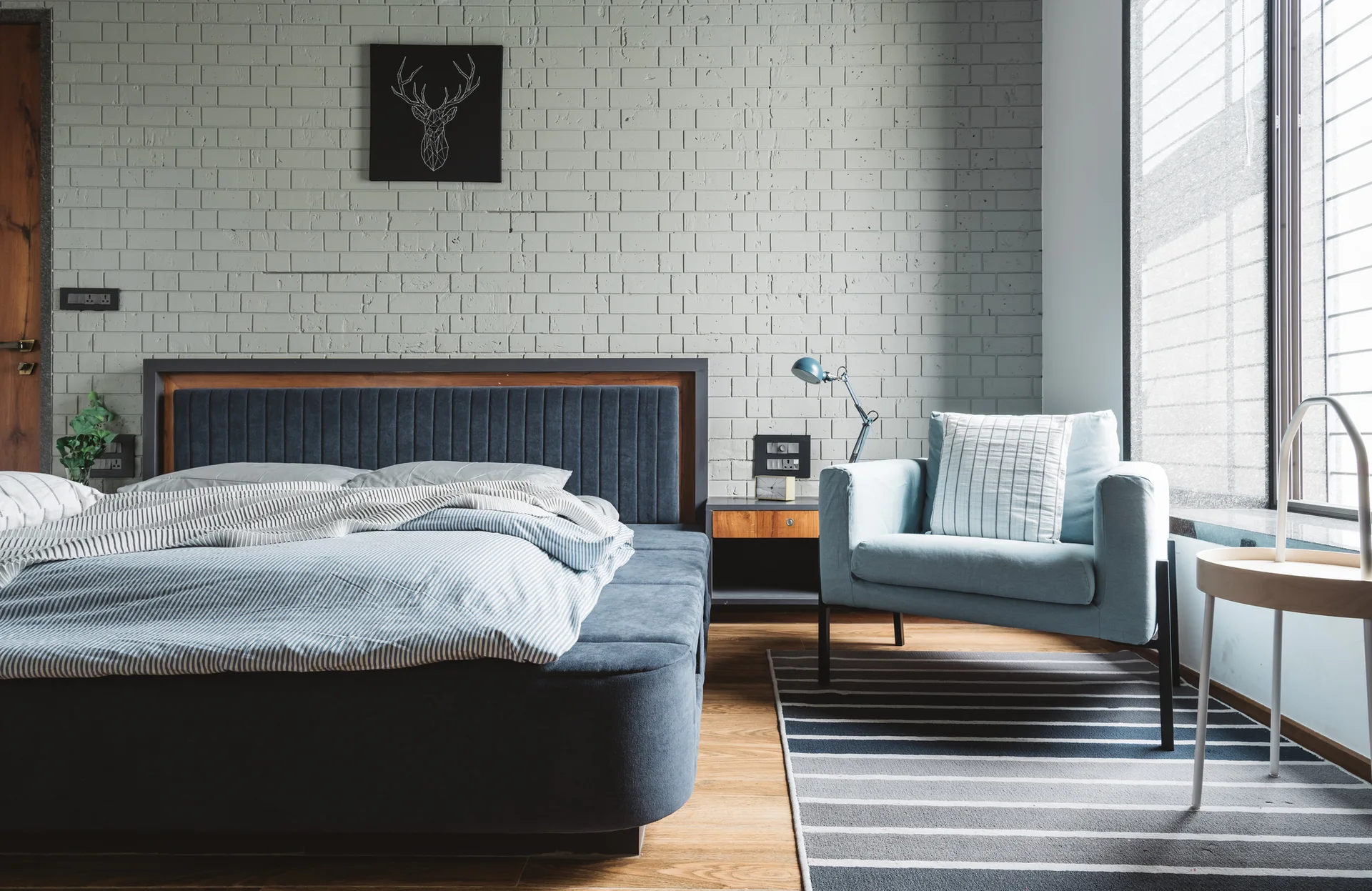 A modern bedroom features a blue upholstered bed and coordinating blue armchair against a light gray painted brick accent wall.
