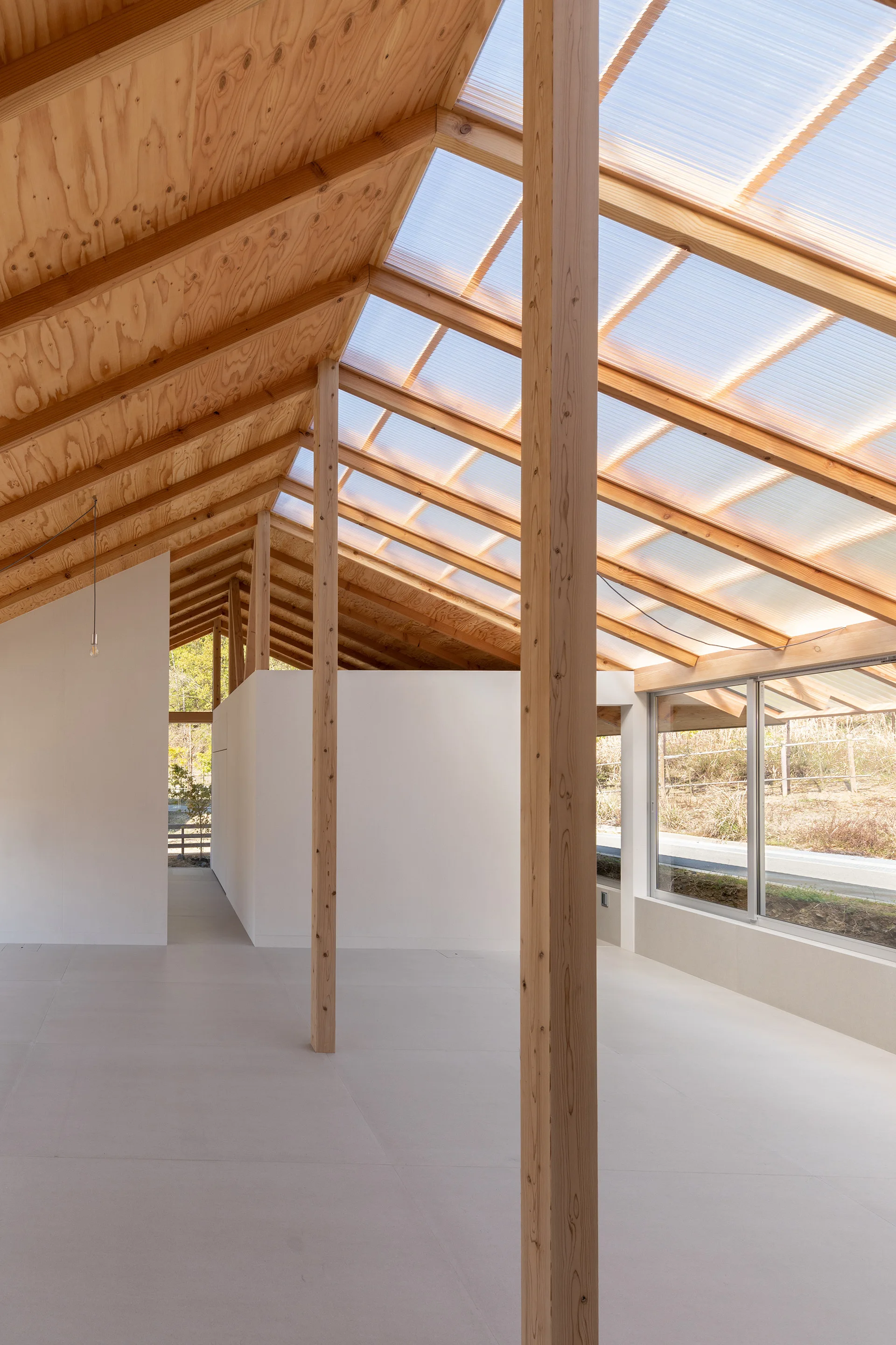 A minimalist interior space features exposed wood rafters, vertical timber columns, white partition walls, and translucent polycarbonate roof panels.