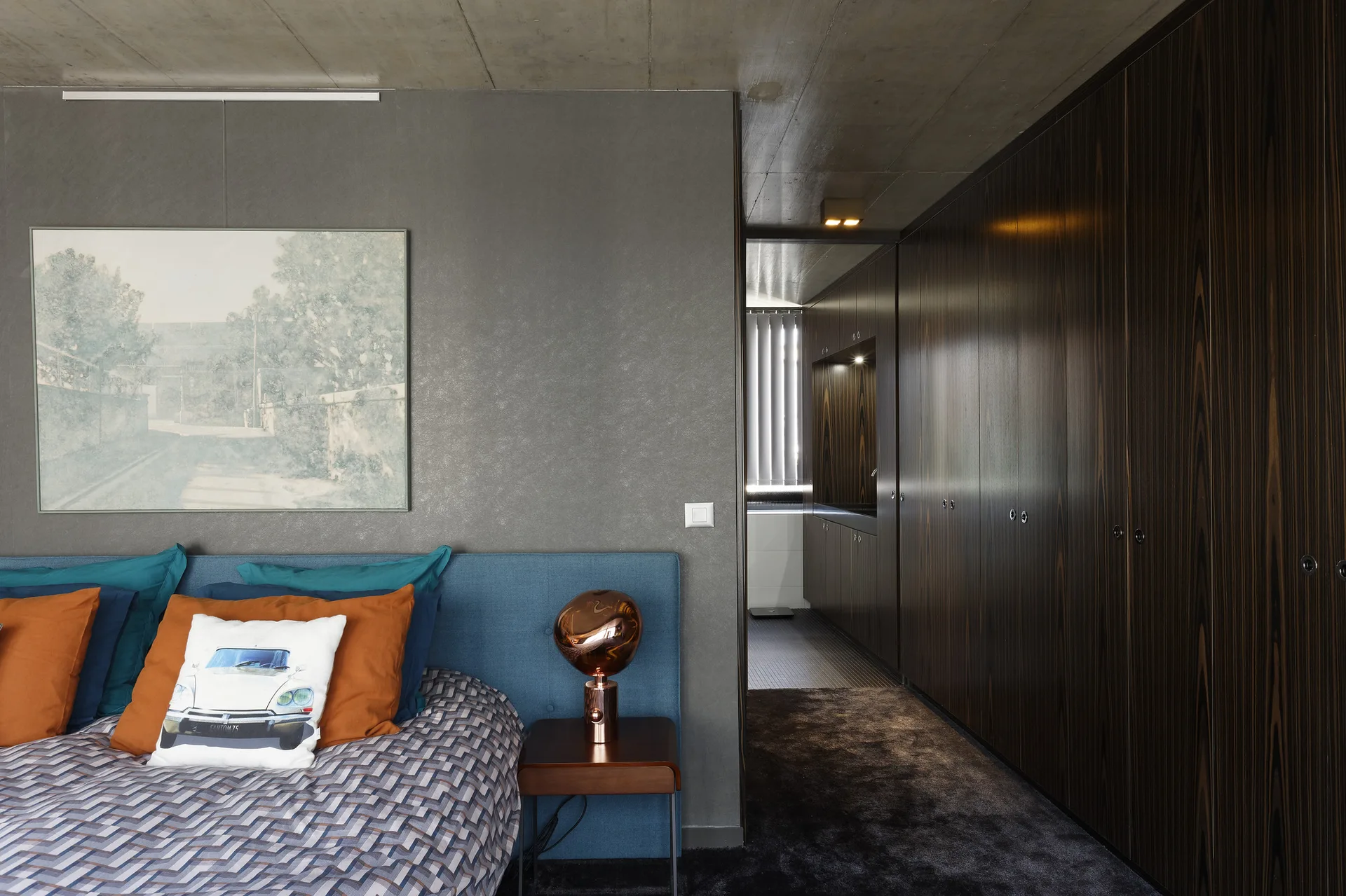 A modern bedroom features a blue upholstered bed set against a grey wall next to a long hallway lined with dark wood closets.