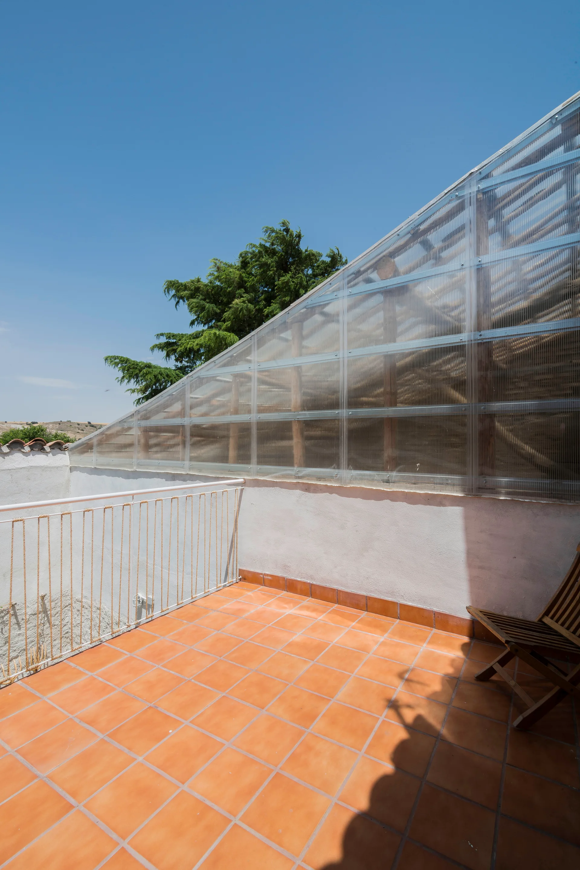 An outdoor rooftop terrace features orange terracotta tile flooring, a white metal and rope railing, and a sloped translucent polycarbonate roof.