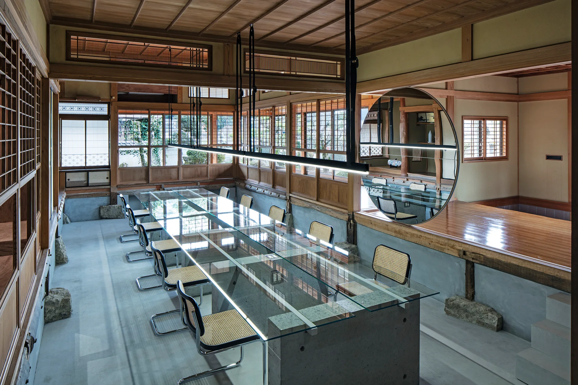 A long glass conference table with cane chairs sits on a concrete floor in a Japanese-style room with wooden latticework and a large circular mirror.
