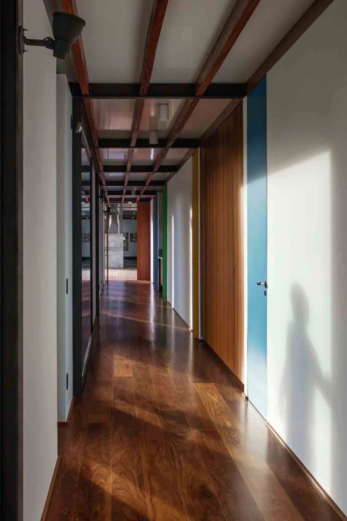 A long hallway features polished dark wood floors, exposed ceiling beams, white walls with blue and wood-toned panels, and a glass wall on the left.