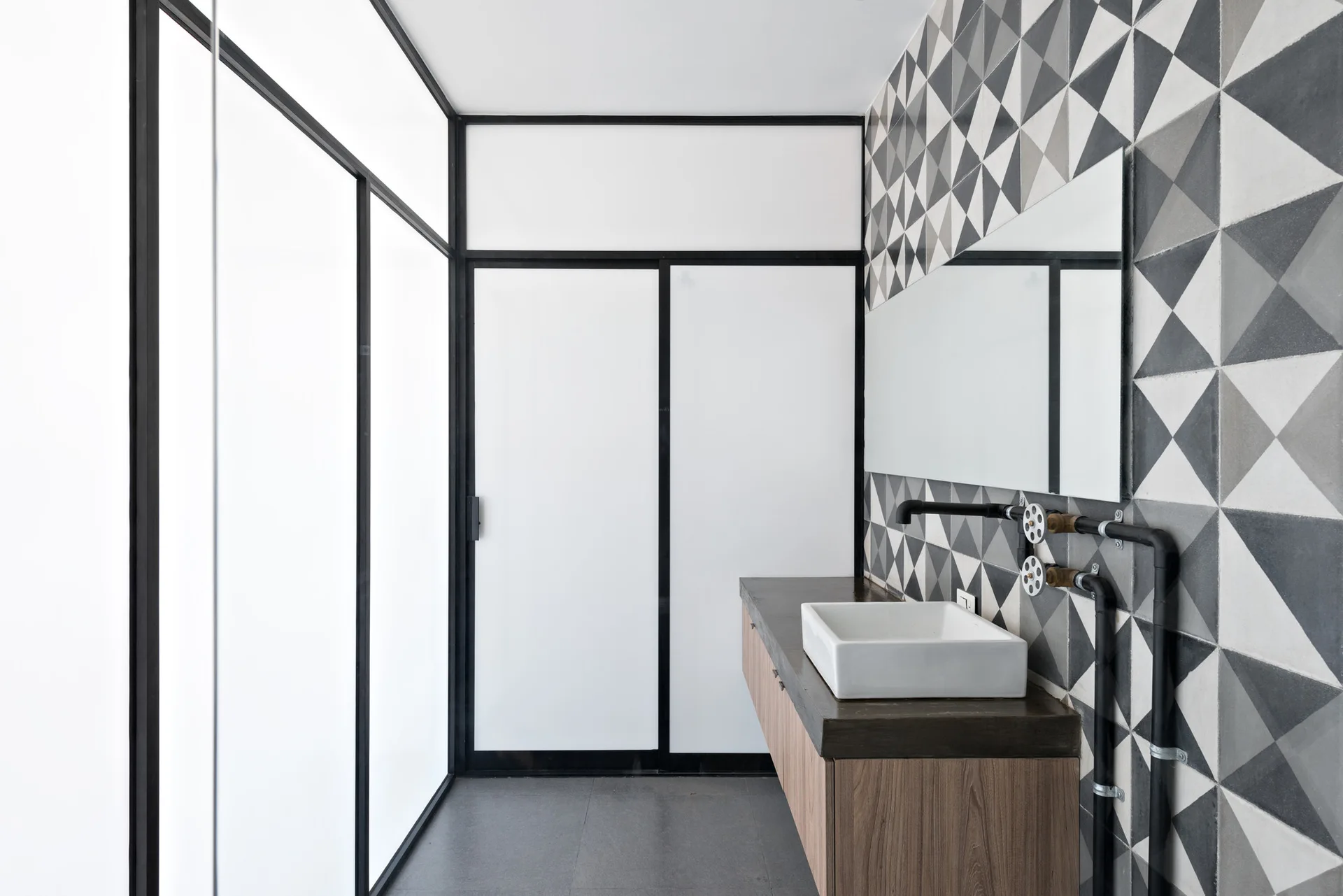 A modern bathroom featuring a wood-toned vanity with a white vessel sink, a geometric tile wall, and black-framed frosted glass partition panels.