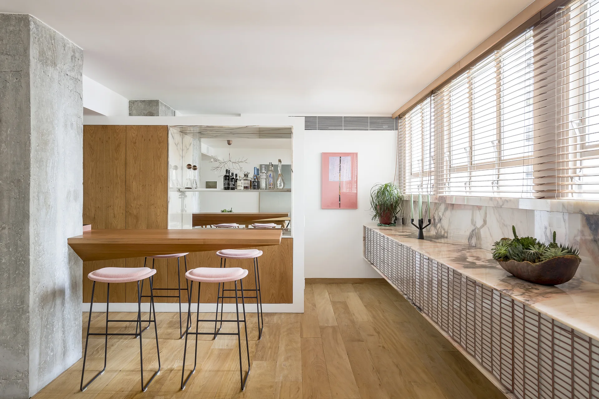 A modern home bar features a wooden counter with pink stools, a raw concrete pillar, and a long stone-topped console beneath a window.