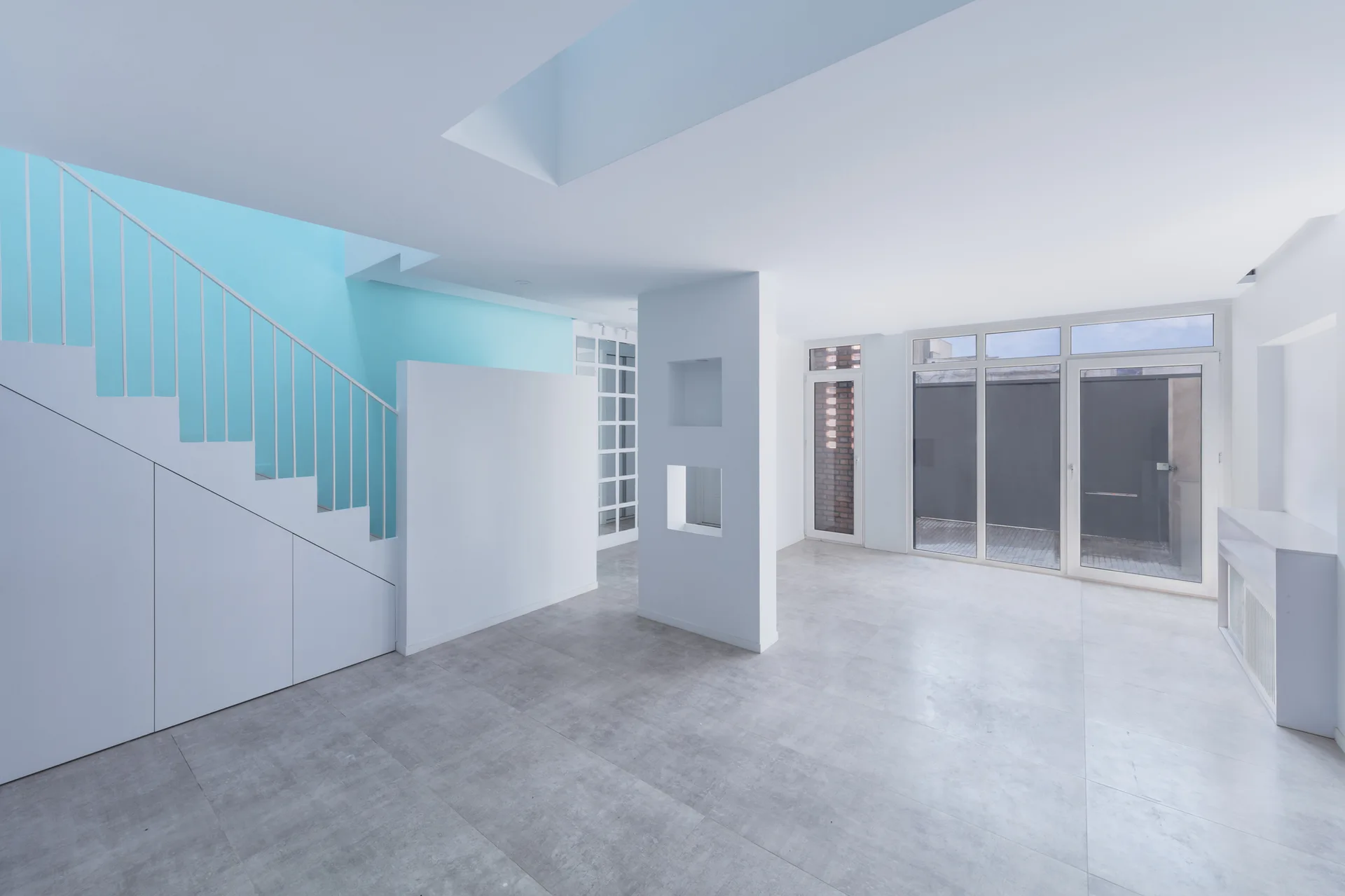 An empty minimalist living room featuring grey tiled flooring, a white staircase with integrated storage cabinets, and a light blue accent wall.