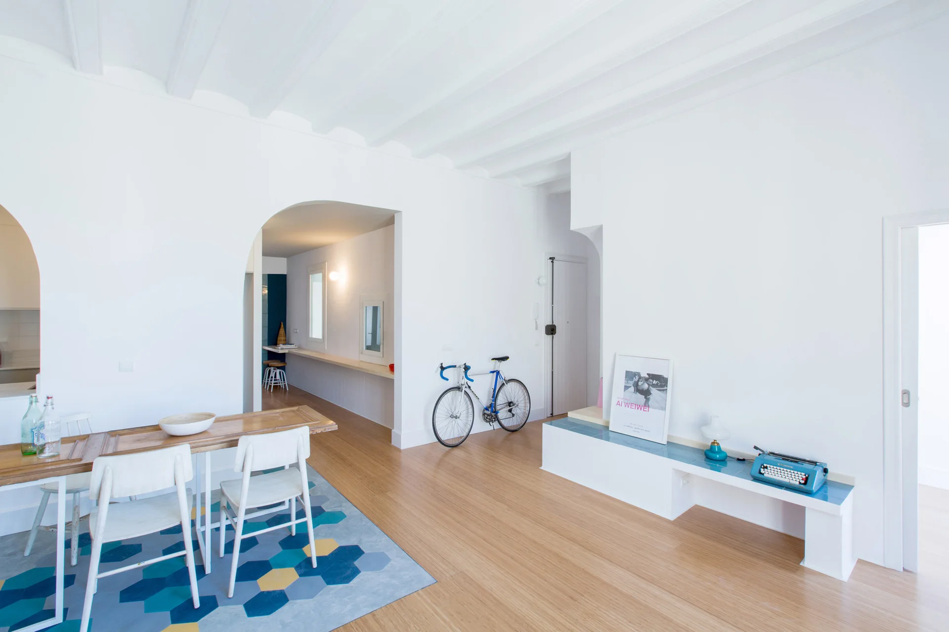 A minimalist white dining room features a wooden table, white chairs on a blue geometric rug, and a low blue-tiled bench.