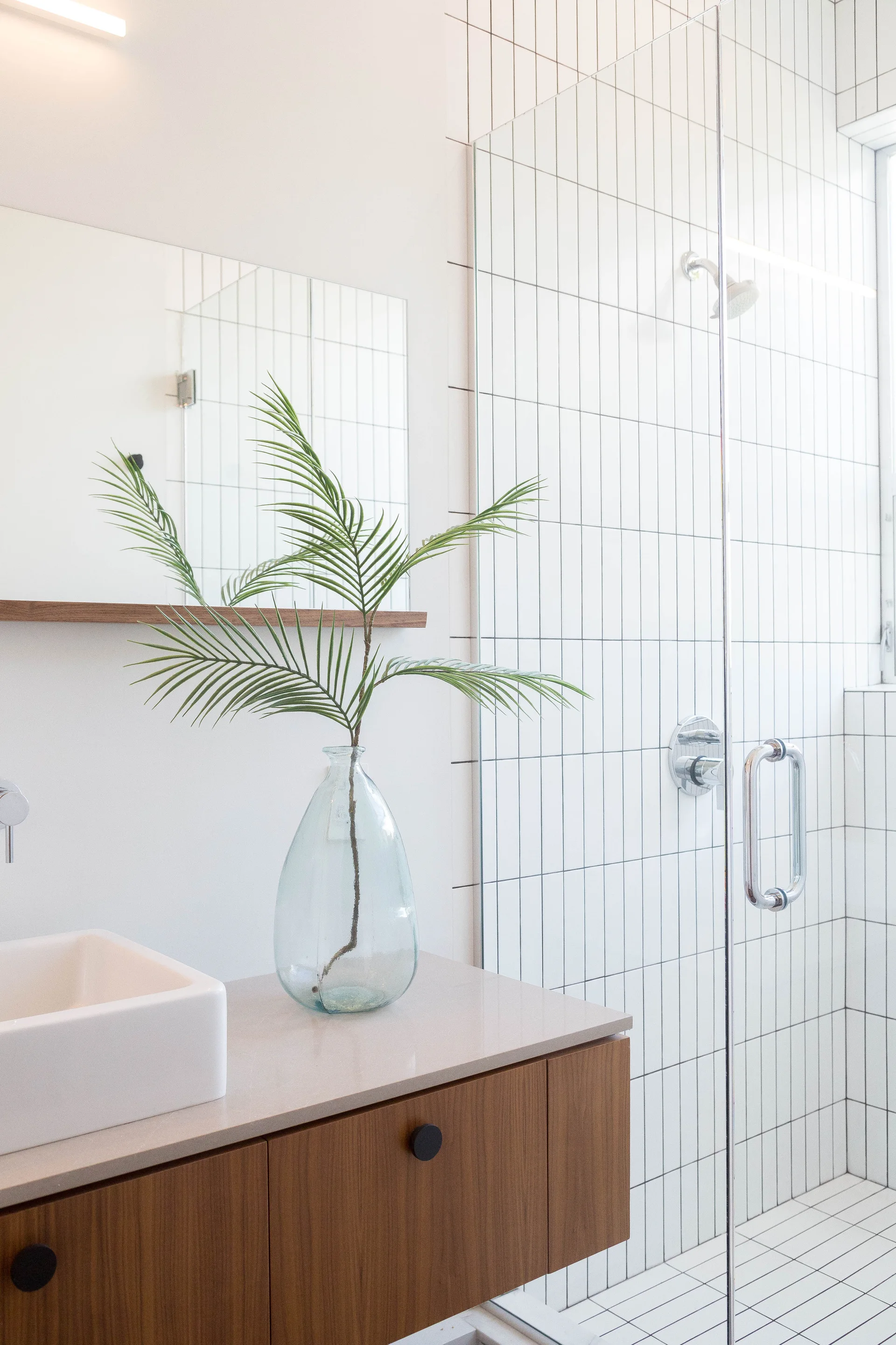 A modern bathroom features a wood floating vanity with a vessel sink next to a glass shower enclosed by vertically stacked white subway tiles.