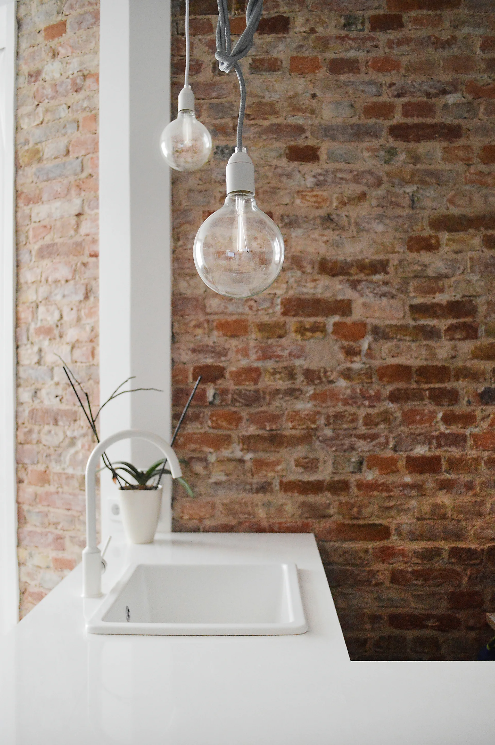 A white kitchen sink and matching arched faucet are installed in a clean white countertop against a rustic exposed red brick wall.