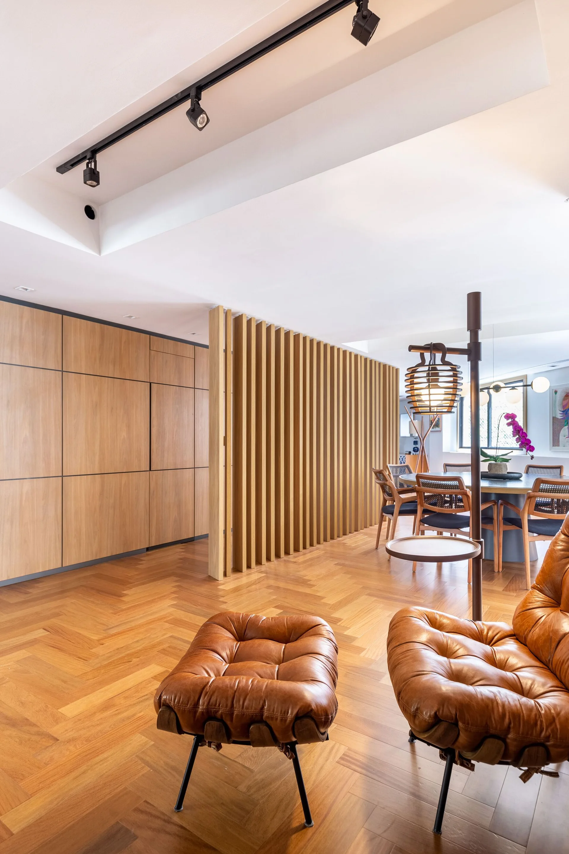 A modern living space features a tufted leather lounge chair, herringbone wood floors, and a vertical wood slat partition screen.