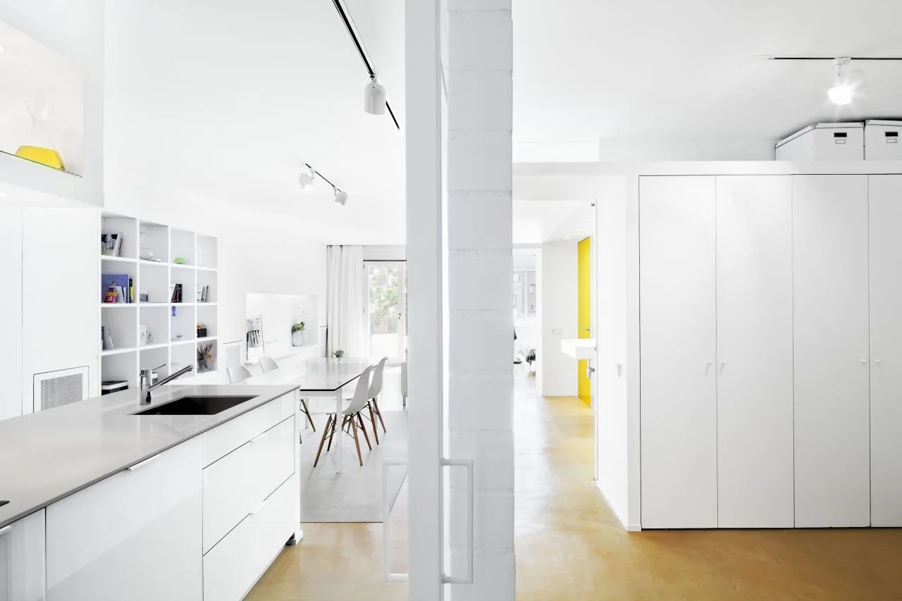A minimalist white kitchen and dining area with a central white island, integrated cabinetry, and light brown poured concrete flooring.
