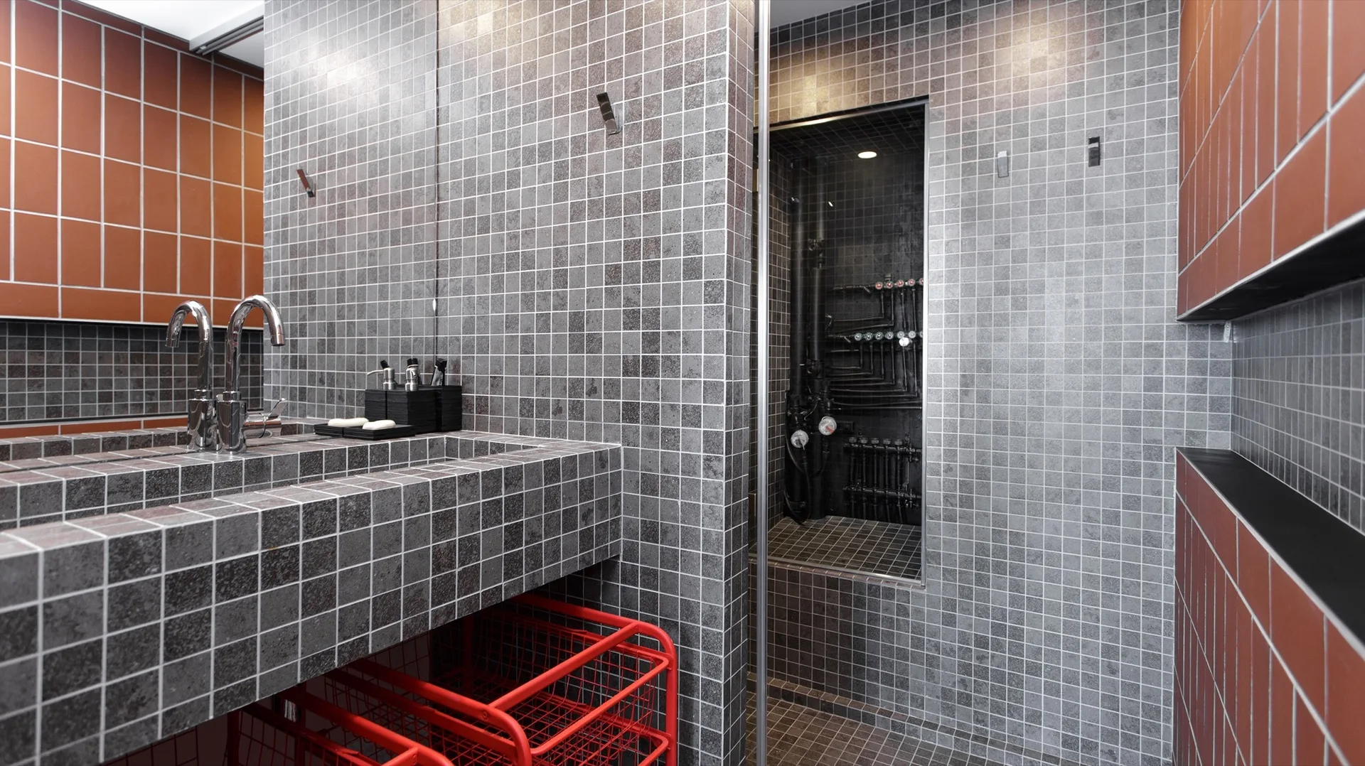 A bathroom featuring grey mosaic tiles on the walls and integrated vanity, red terracotta tiles, a chrome faucet, and a niche showing black plumbing pipes.