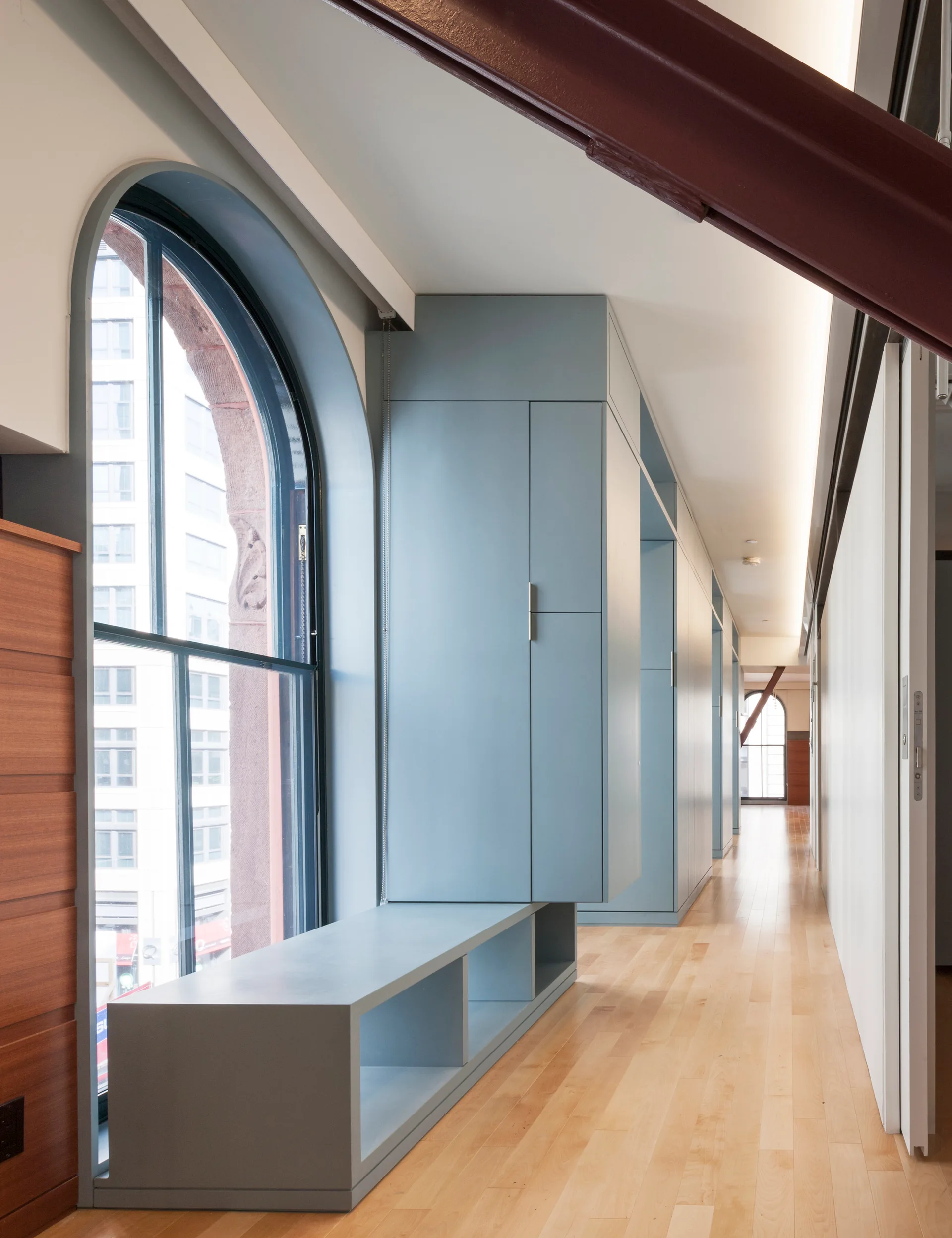 A long hallway features built-in muted blue cabinets with an integrated window bench alongside a historic arched window and light wood floors.
