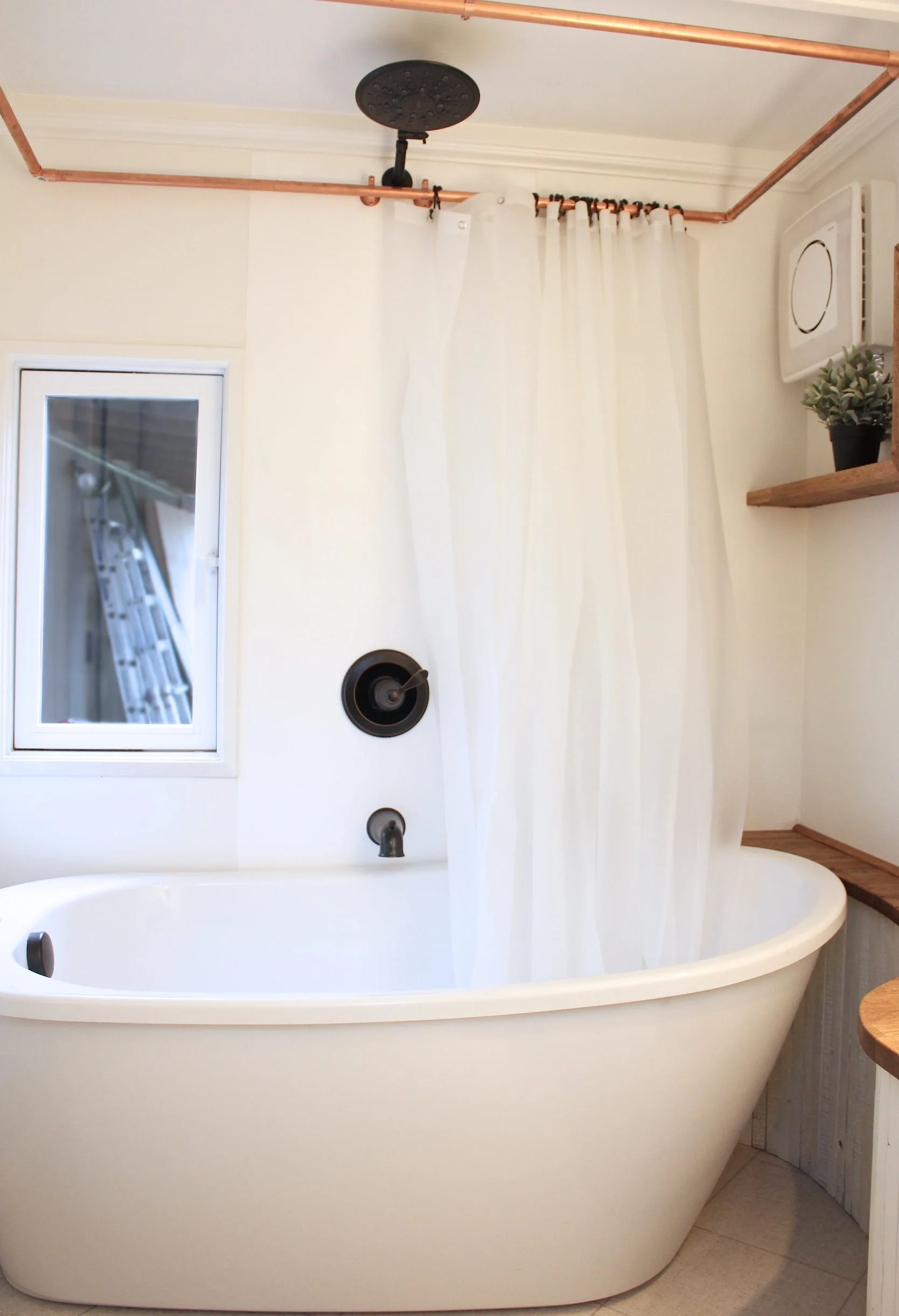 A white freestanding oval bathtub features a translucent shower curtain hanging from exposed copper piping and black matte hardware.