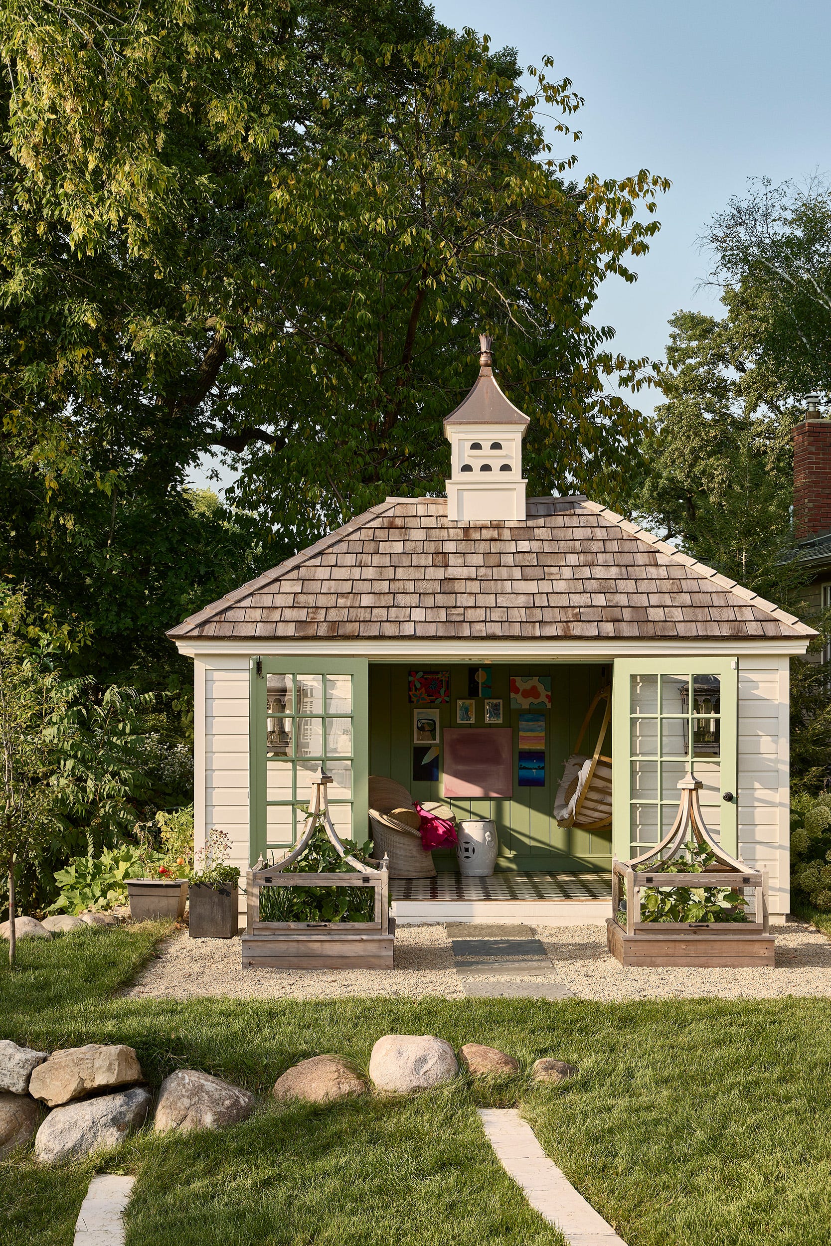 A backyard garden shed with open sage green french doors, a shingled roof with a cupola, and two wooden planters in front.