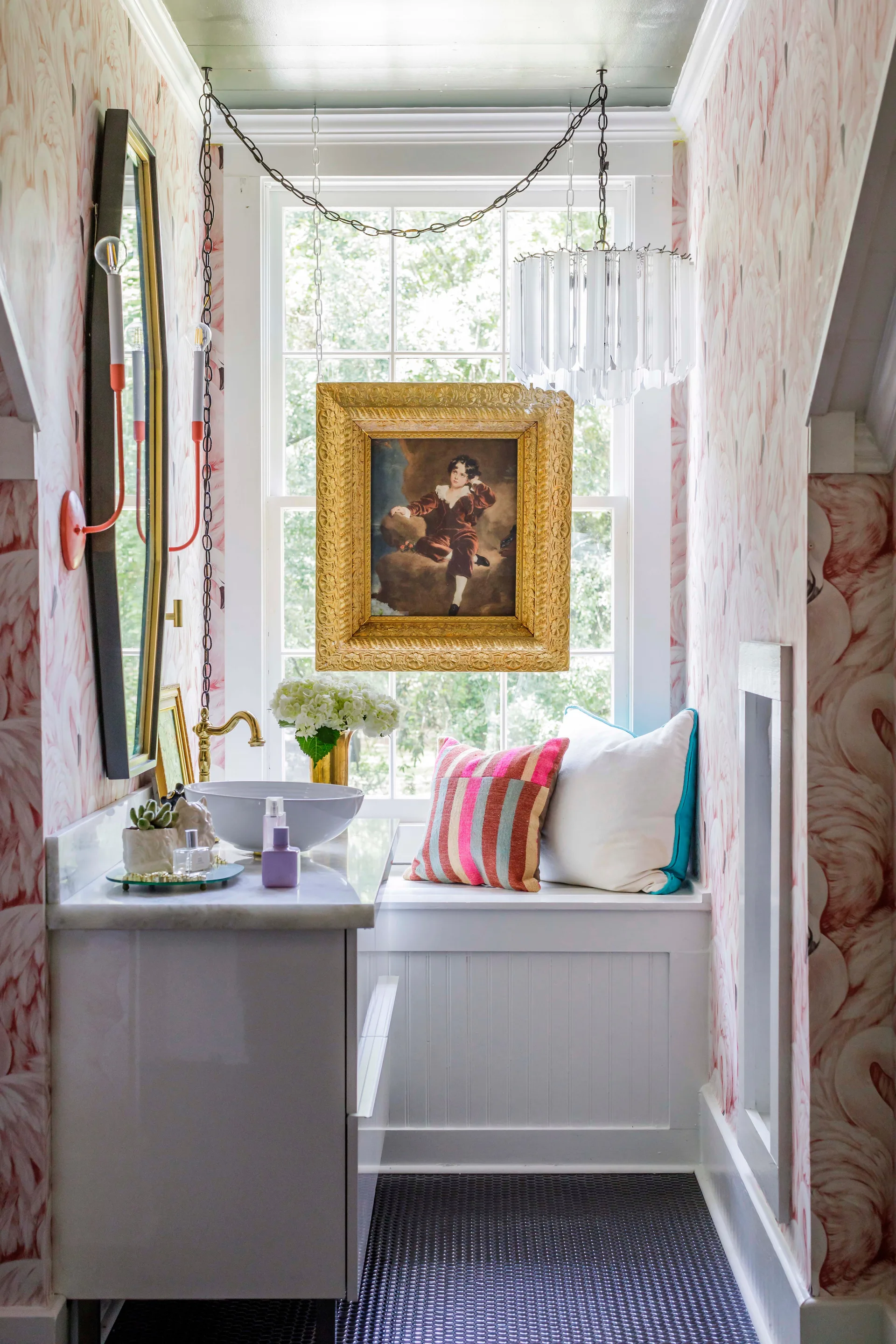A narrow bathroom features pink flamingo wallpaper, a white vanity with a vessel sink, and a built-in window seat beneath a suspended framed portrait.