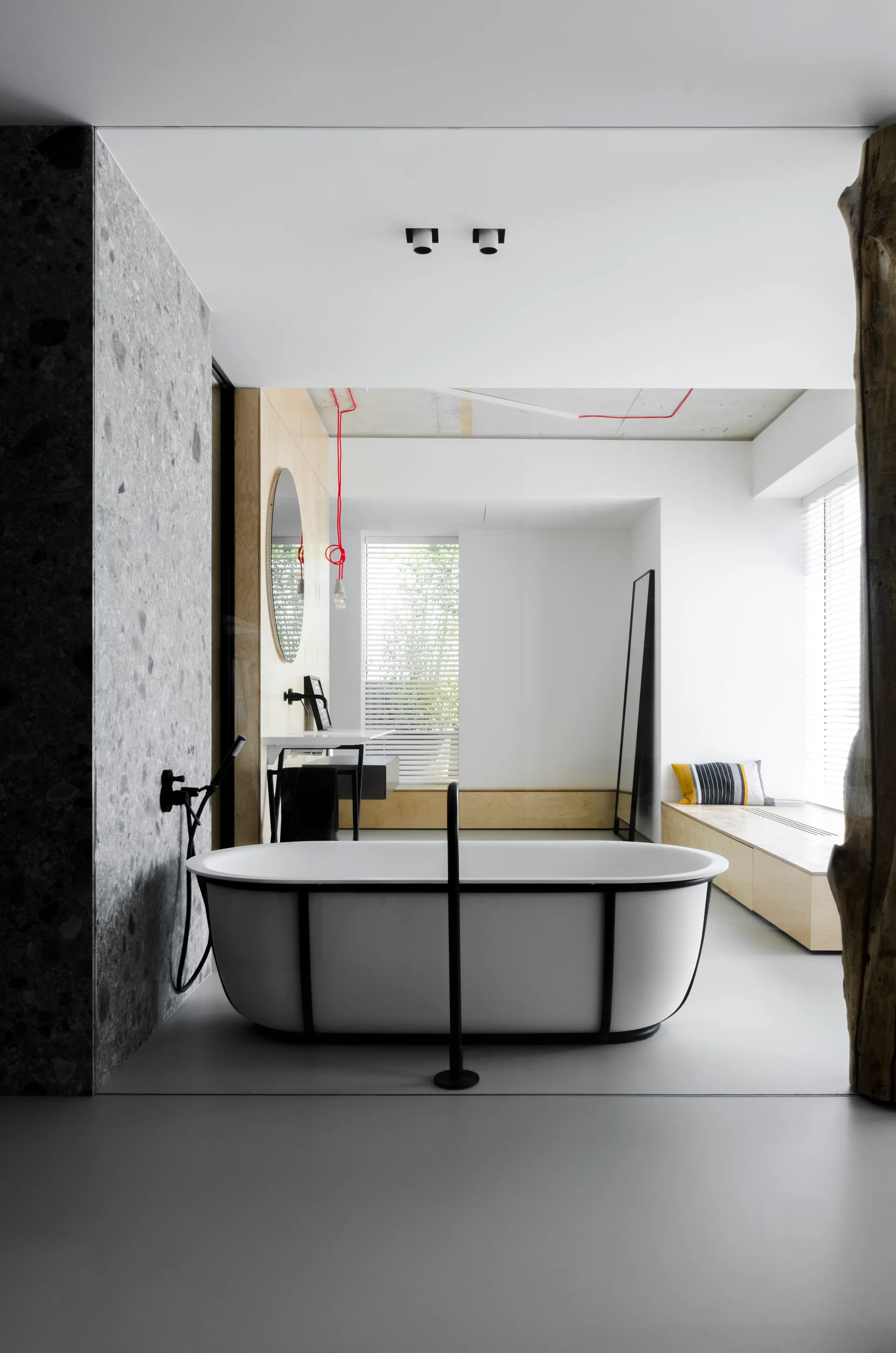 A modern minimalist bathroom with a black-framed freestanding white bathtub, stone wall tile, and a built-in plywood window bench.