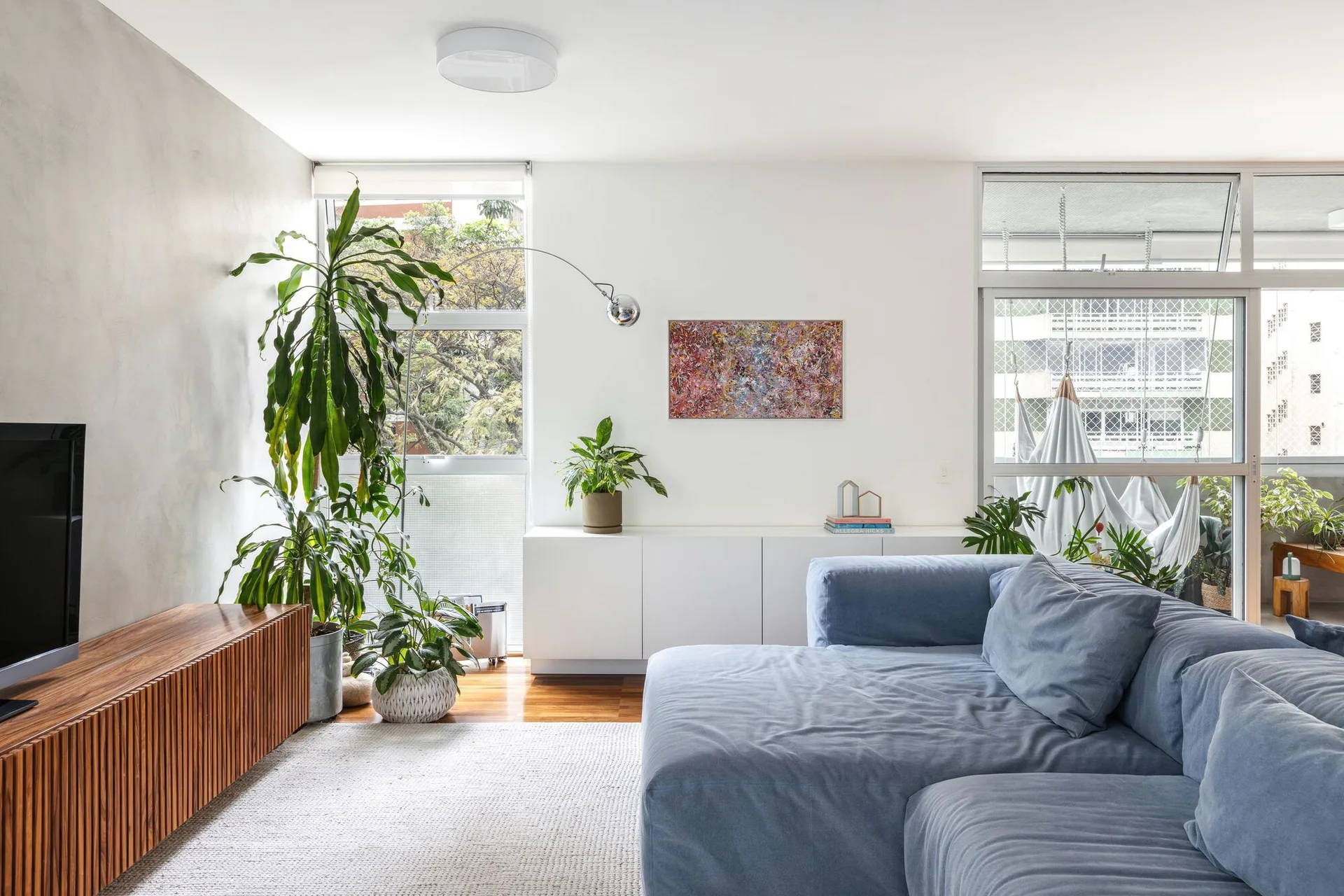 A bright living room featuring a blue sectional sofa, a slatted wood media console, potted plants, and a balcony window.