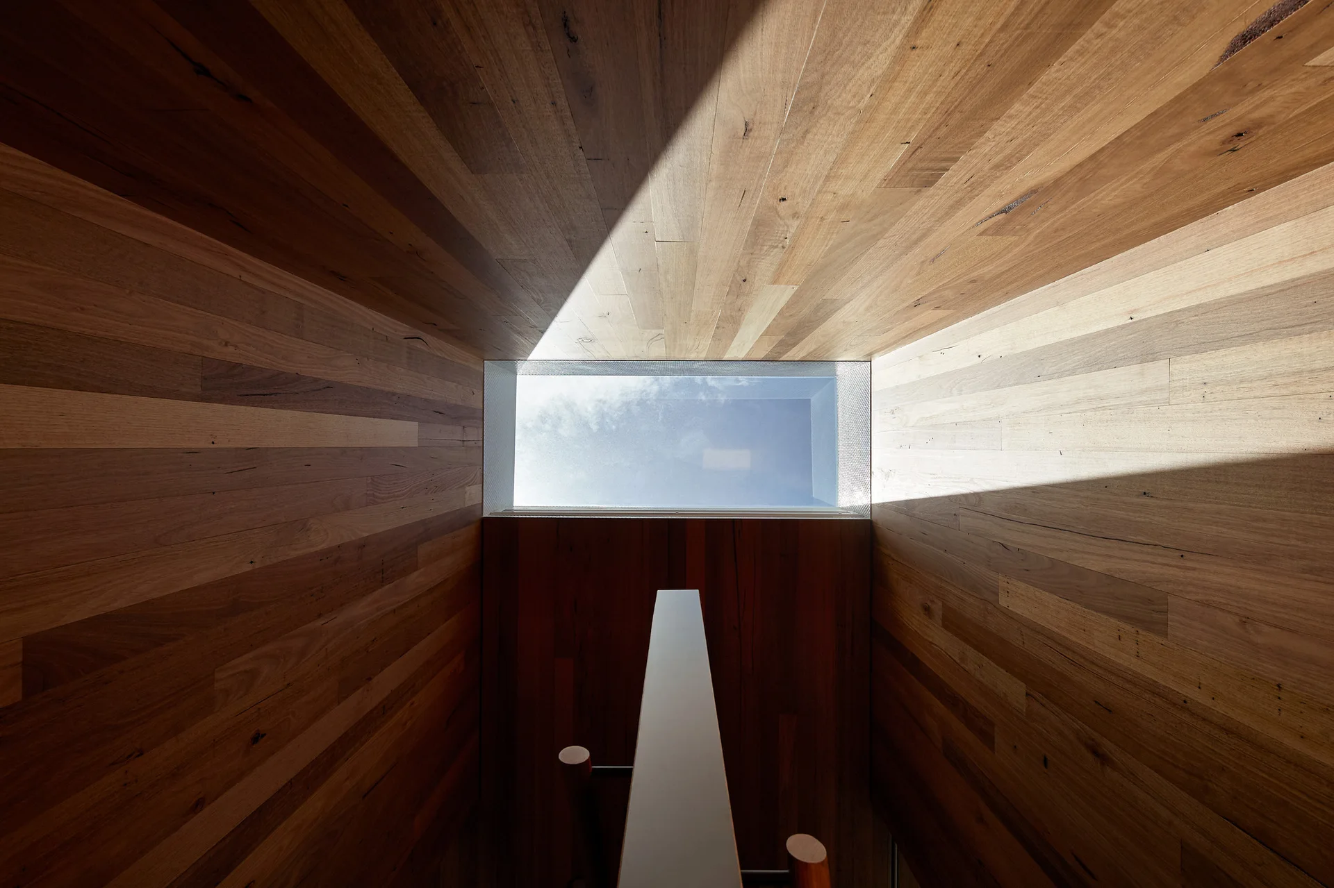 Looking upward through a wooden-paneled stairwell toward a rectangular skylight showing a blue sky with light clouds.