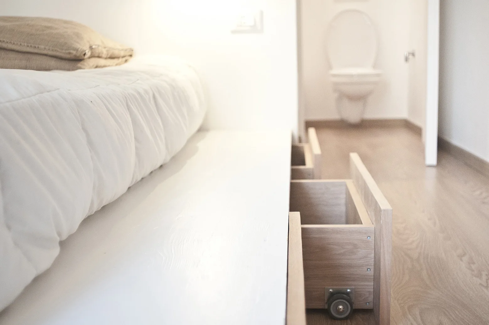 A white platform bed features wooden under-bed storage drawers on casters, with a bathroom visible in the background.